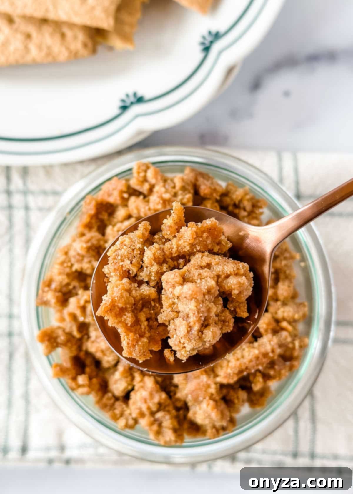 No-Bake Graham Cheesecake Delights 3 An overhead close-up photo of golden-brown graham cracker crumbles resting on a bronze spoon, positioned over a glass jar, highlighting their delicious texture.