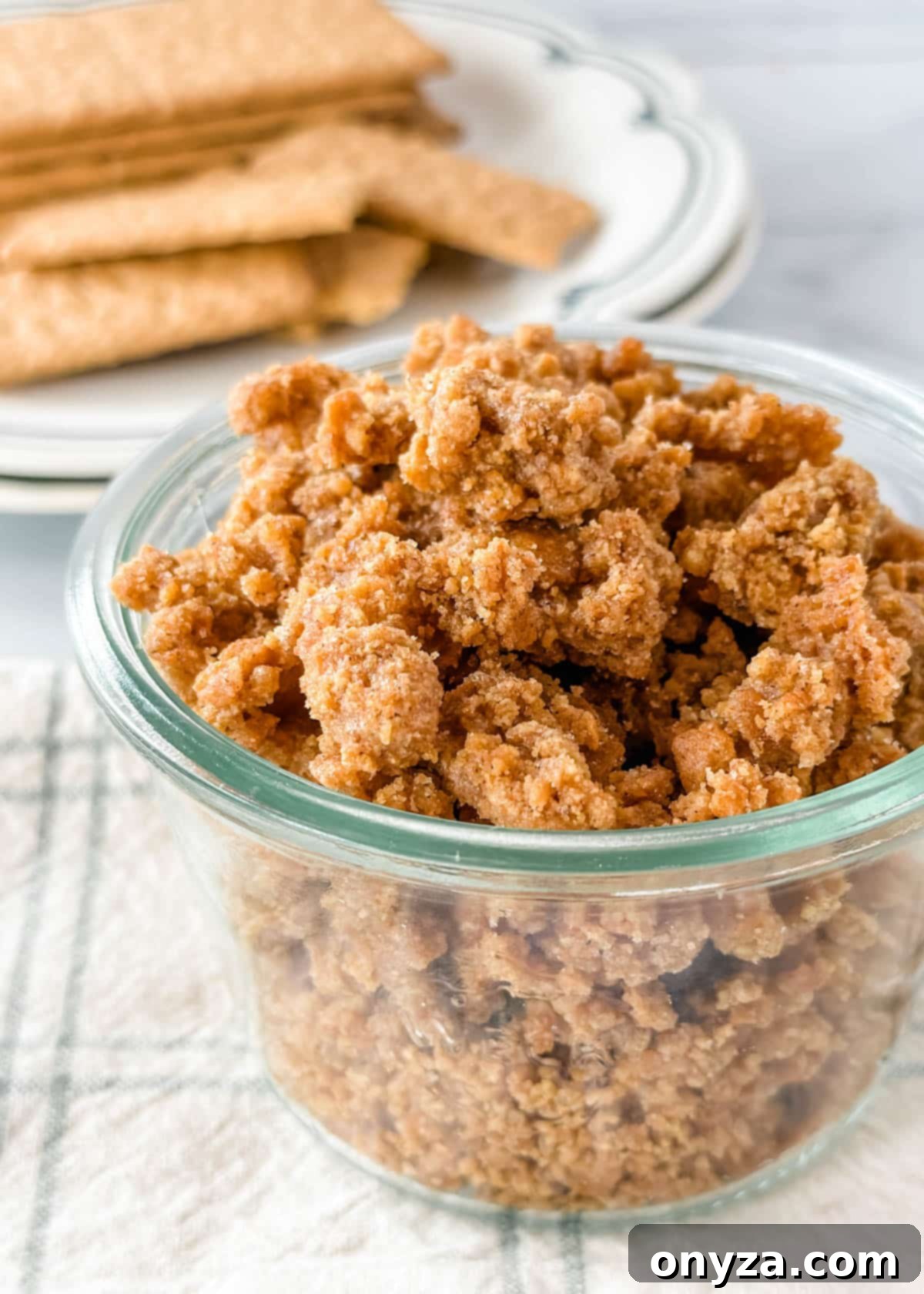 Homemade graham cracker crumbles in a glass jar, ready to be served as a crunchy dessert topping, resting on a white and green checkered napkin with whole graham crackers blurred in the background.