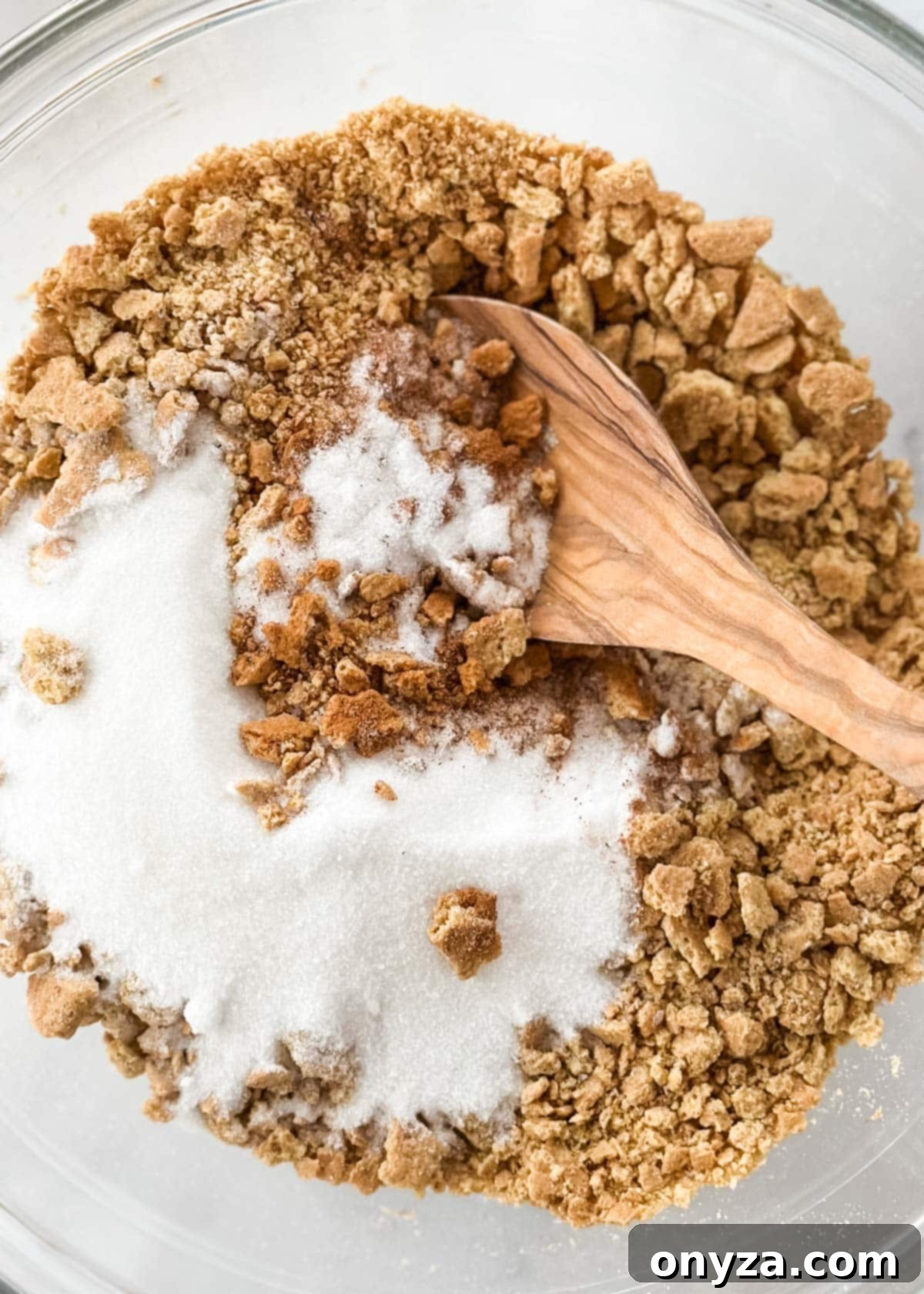 Closeup of graham cracker crumbs, sugar, and cinnamon in a glass bowl, ready for mixing, with a wooden spoon resting nearby.