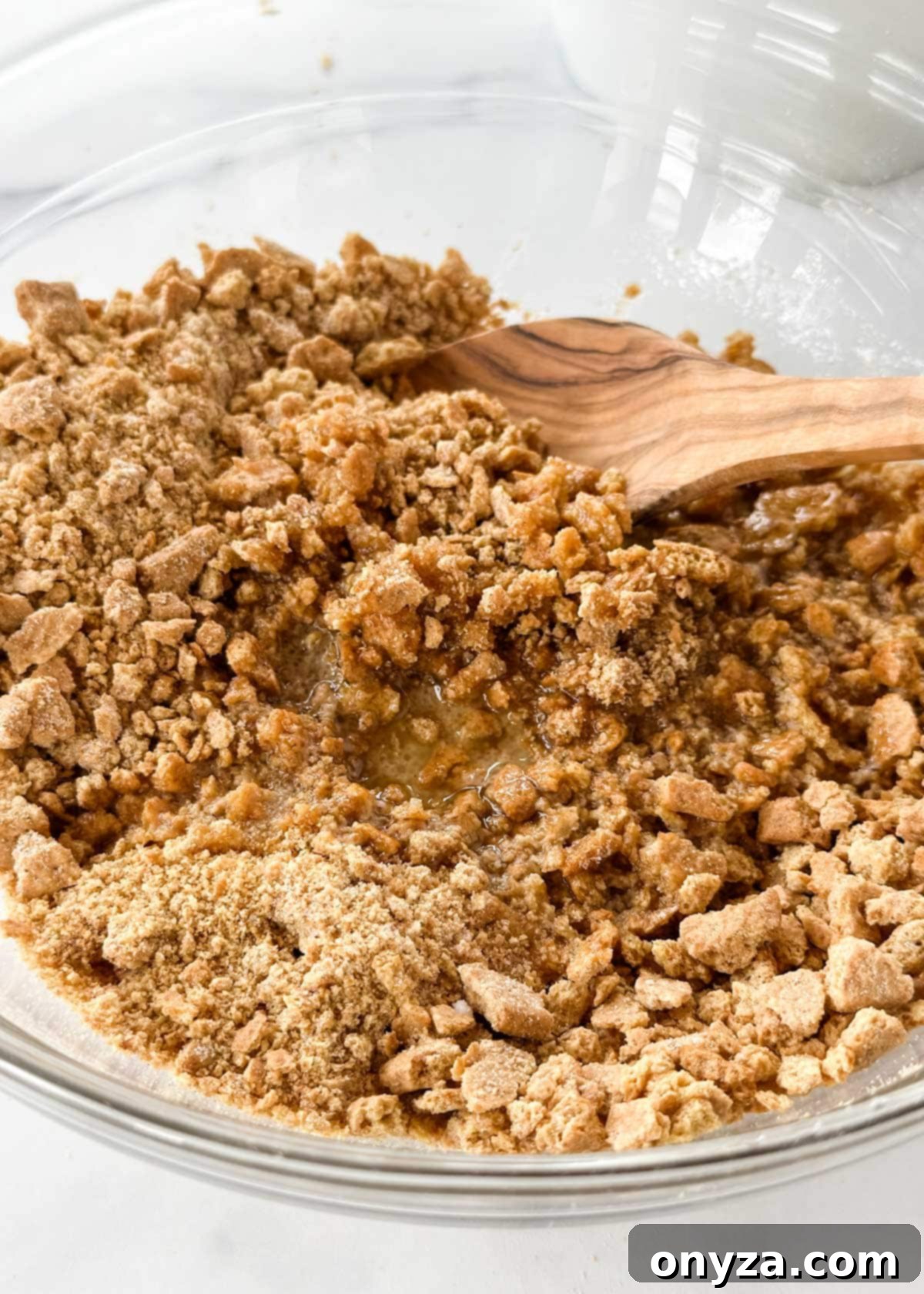 Mixed graham cracker crumbs, sugar, and melted butter in a clear glass bowl, being stirred with a wooden spoon, ready for baking.