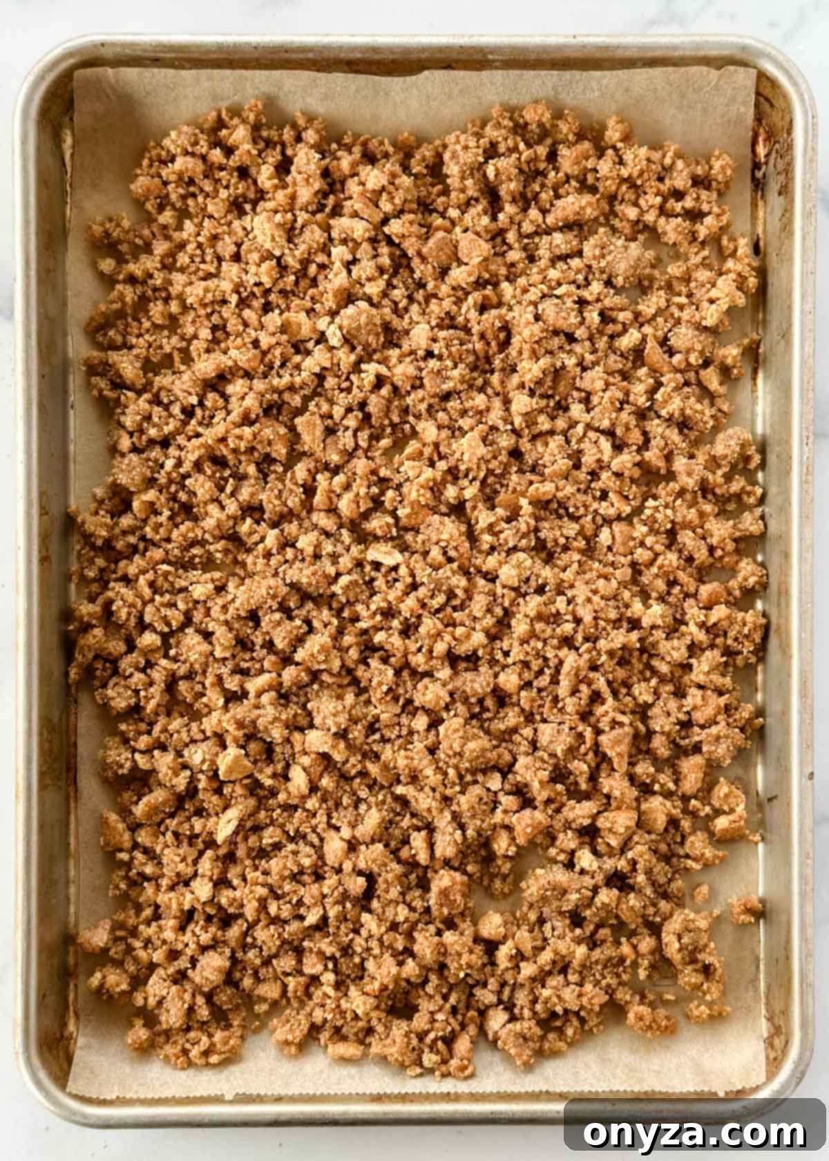 Unbaked graham cracker crumbles spread evenly on a parchment-lined baking sheet, ready to be toasted in the oven.