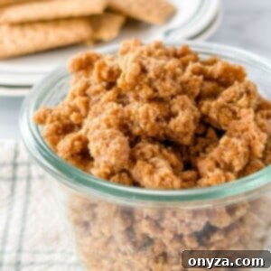Homemade graham cracker crumbles in a glass jar, ready to be served as a crunchy dessert topping, resting on a white and green checkered napkin with whole graham crackers blurred in the background.