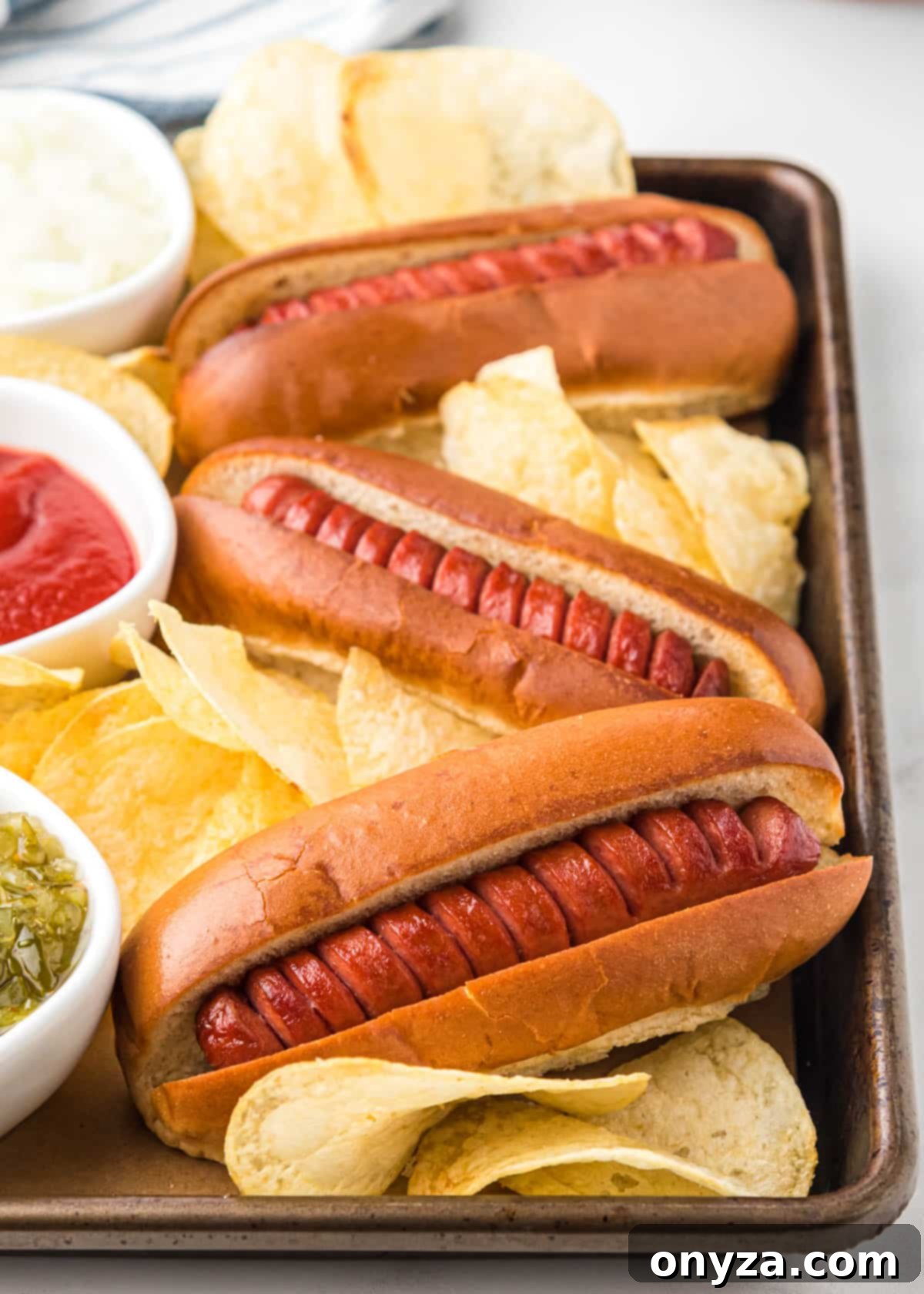 Perfectly cooked air fryer hot dogs in toasted buns, served on a baking sheet with potato chips and bowls of classic condiments like relish, ketchup, and chopped onions.
