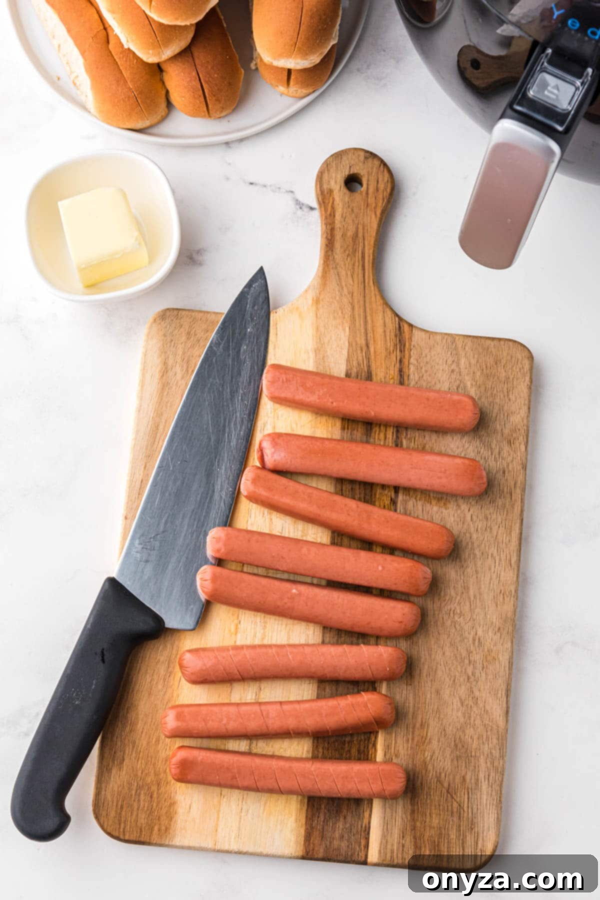 Overhead shot of hot dogs arranged on a rustic wooden board next to a chef's knife, showcasing the preparation stage before air frying.