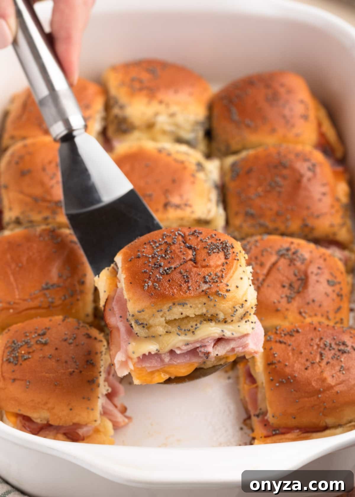 baked ham and cheese slider being removed from a white baking dish with a stainless steel server