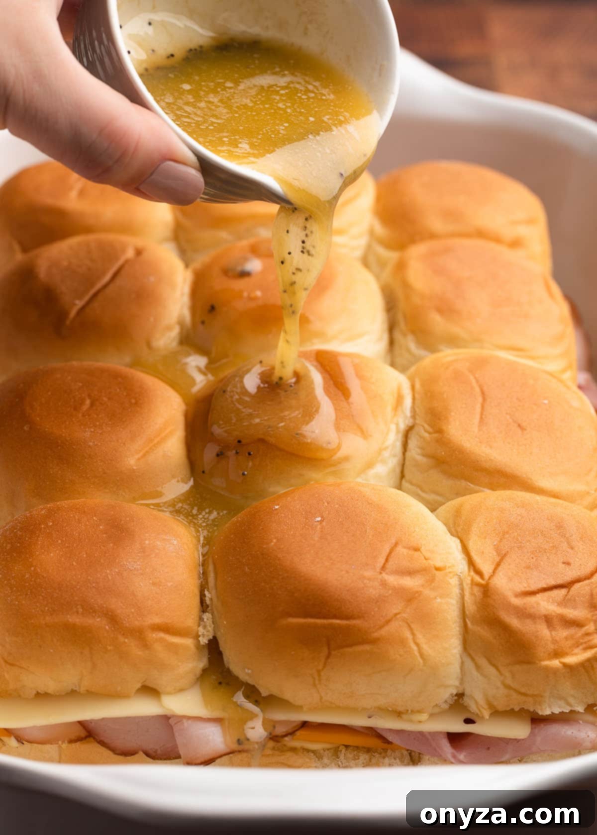 butter sauce being poured over unbaked ham and cheese sliders in a white ceramic baking dish