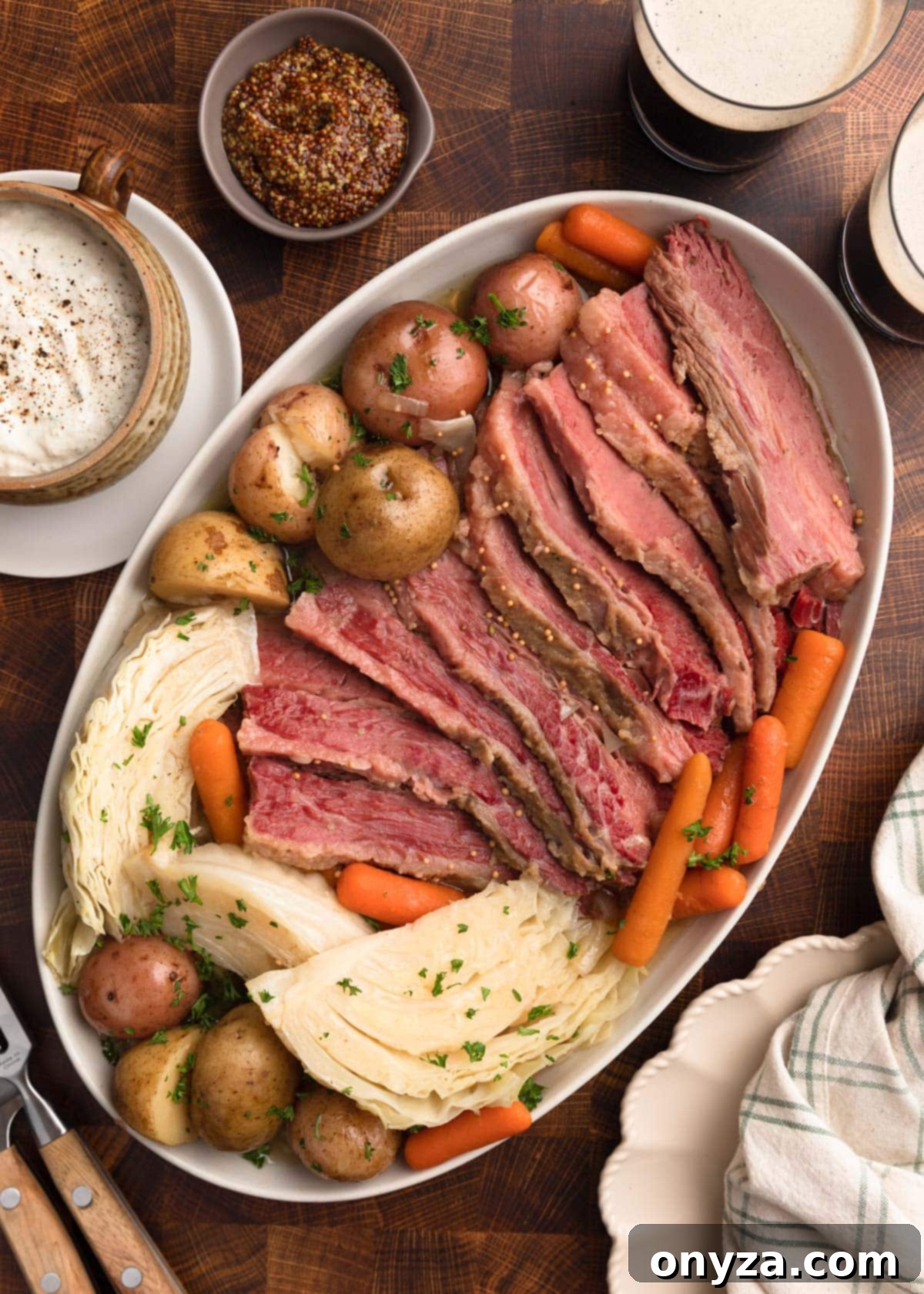 Effortless Slow Cooker Corned Beef and Cabbage 12 Overhead photo of perfectly cooked, sliced corned beef presented on a platter with baby potatoes, baby carrots, and tender cabbage wedges. Accompaniments include bowls of grainy mustard and horseradish sauce, alongside two glasses of stout beer.
