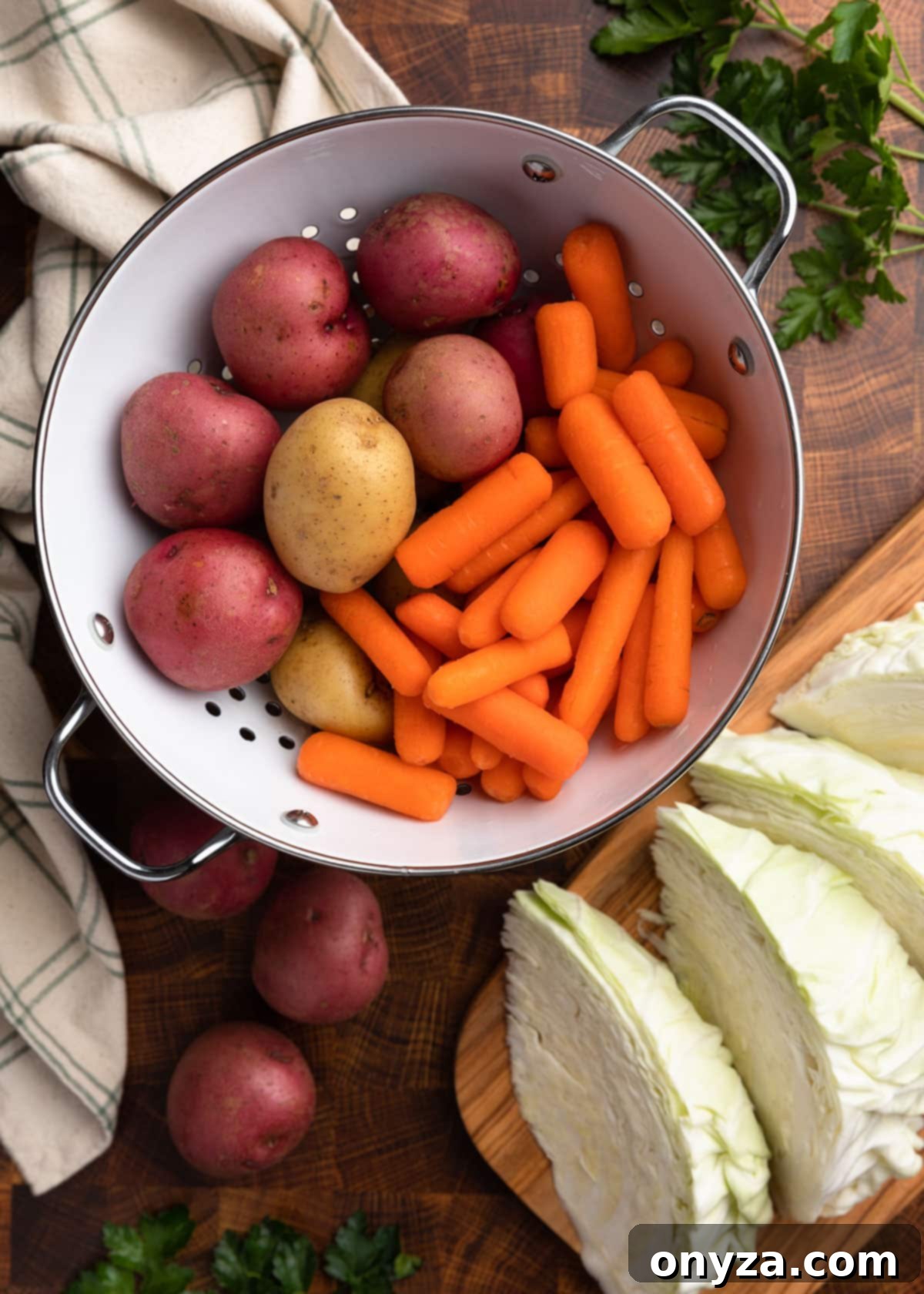Effortless Slow Cooker Corned Beef and Cabbage 9 Overhead photo of vibrant baby red and yellow potatoes, and baby carrots in a white colander next to a cutting board with freshly cut cabbage wedges.