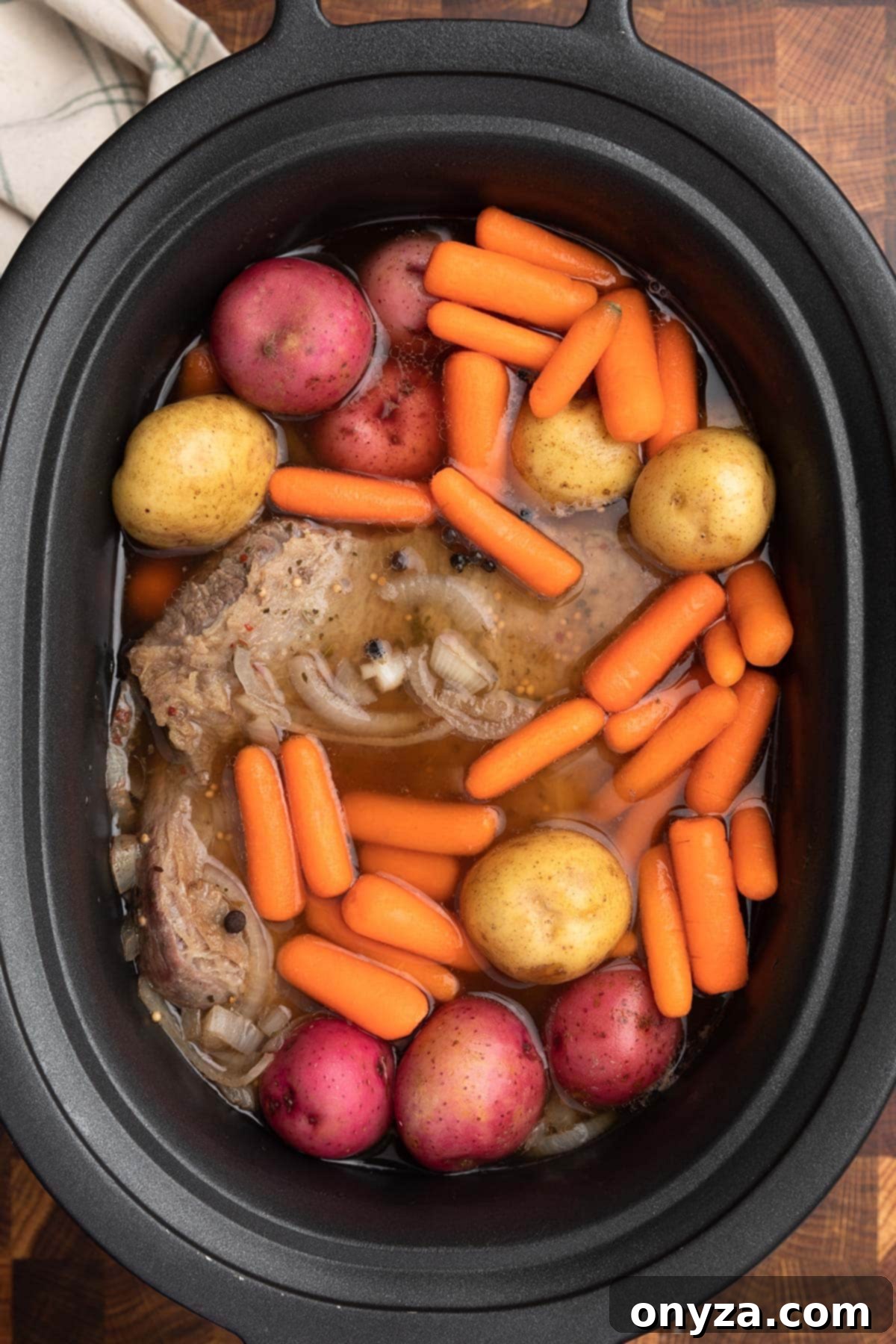 Effortless Slow Cooker Corned Beef and Cabbage 10 Overhead photo of corned beef in a slow cooker insert with raw baby potatoes and carrots added after the initial cooking period.