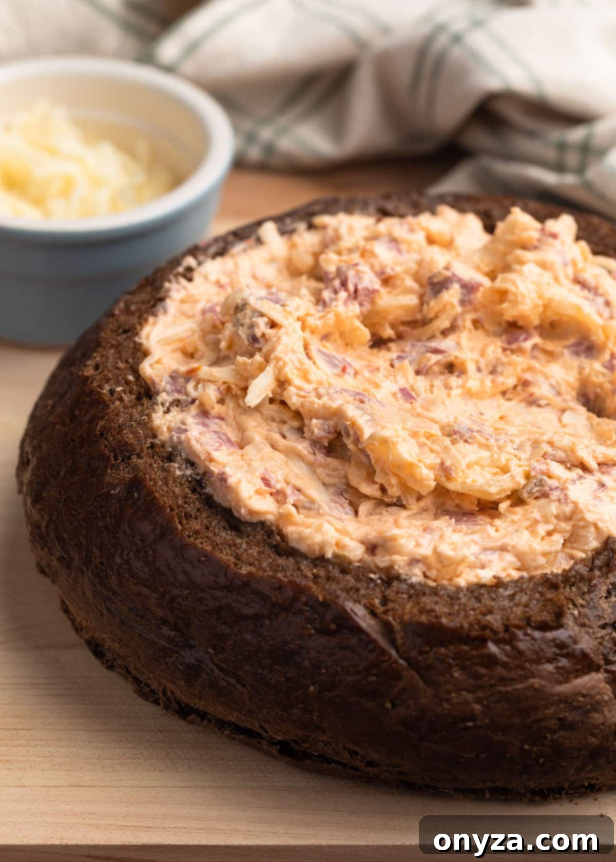 Irresistible Reuben Dip 7 unbaked reuben dip in a pumpernickel bread bowl on a light wood cutting board with a bowl of shredded swiss cheese on the side