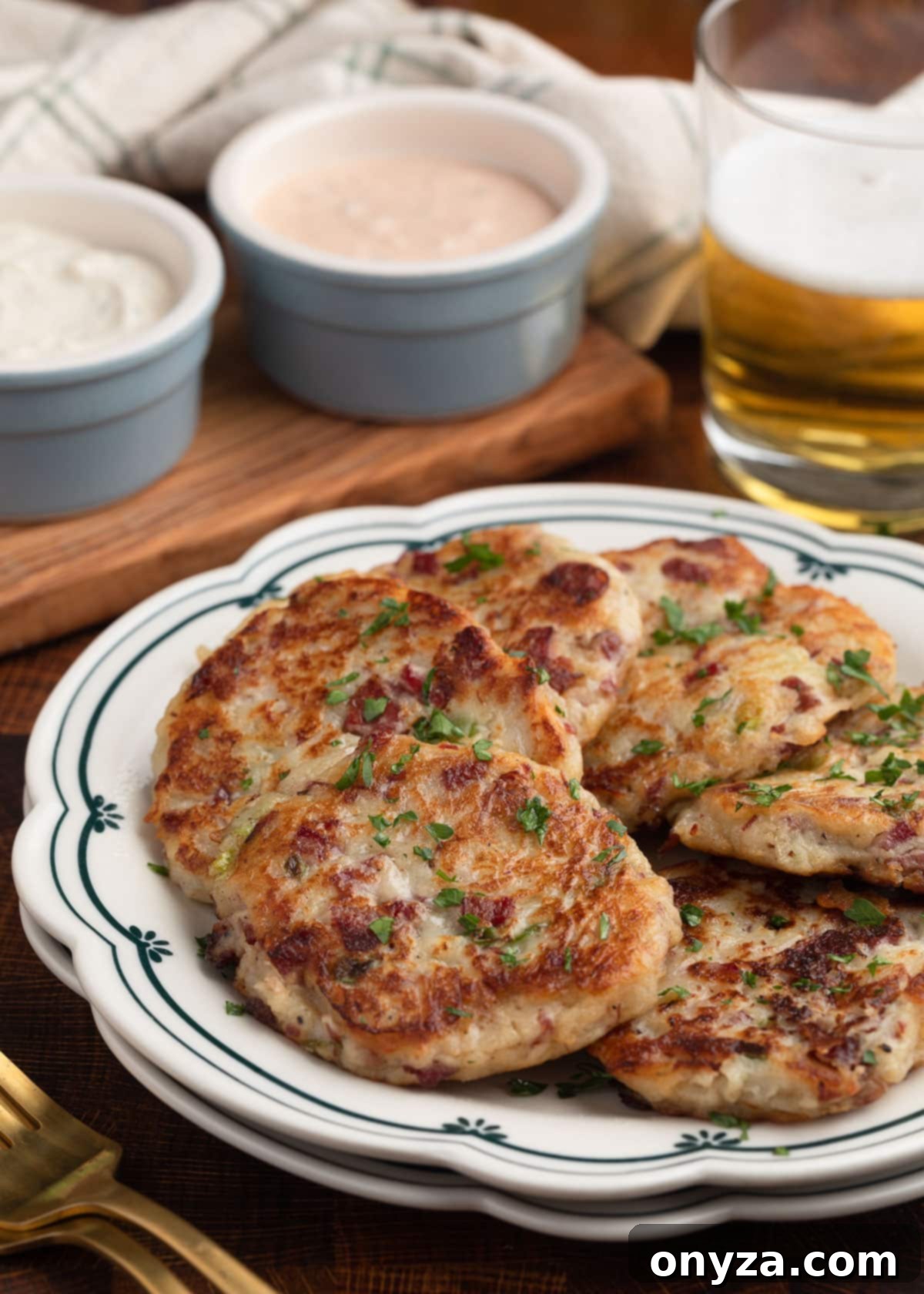 Golden Corned Beef Potato Fritters 2 Crispy Corned Beef and Potato Cakes served on a decorative white and blue scalloped dish, with a glass of beer, horseradish sauce, and Russian dressing in the background, ready to be enjoyed.