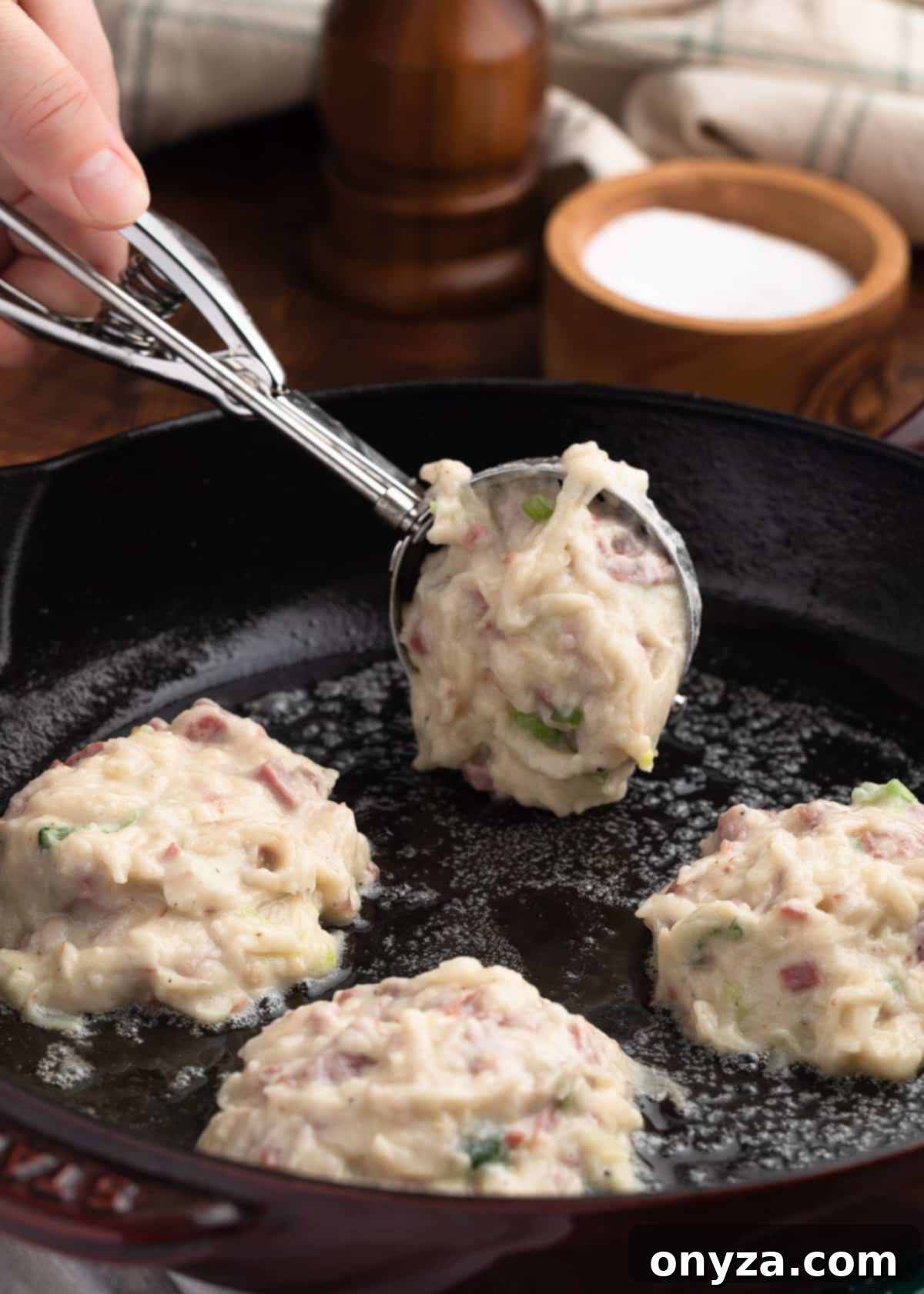Golden Corned Beef Potato Fritters 7 A stainless steel ice cream scoop is used to carefully drop portions of corned beef and potato cake batter into a hot cast iron frying pan, forming small, uniform pancakes, with space left between each for optimal frying.