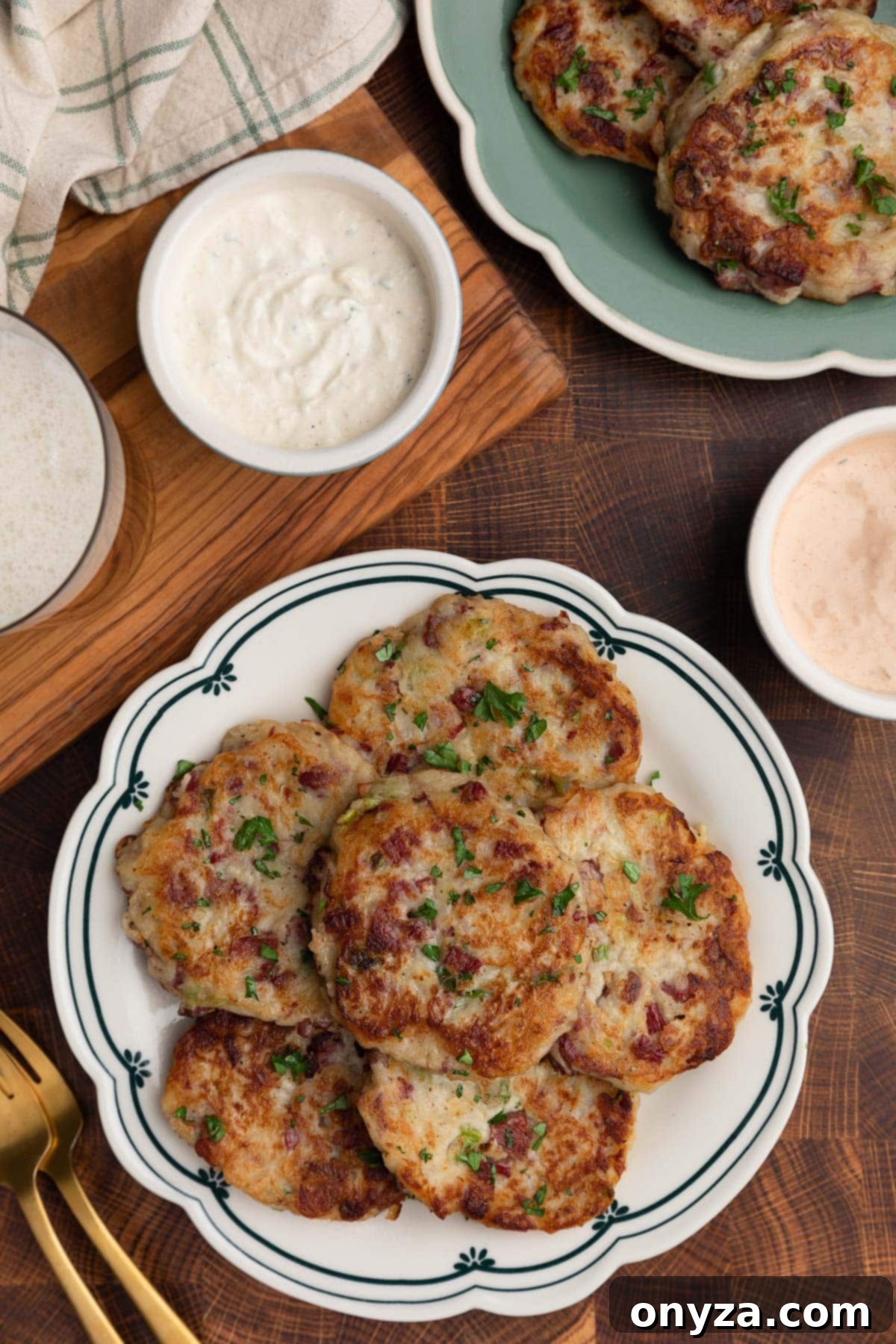 Golden Corned Beef Potato Fritters 9 An overhead view of golden-brown corned beef and potato cakes elegantly arranged on a white and blue scalloped dish, accompanied by small bowls of Creamy Horseradish Sauce and Russian Dressing, set on a rustic wooden butcher block surface.
