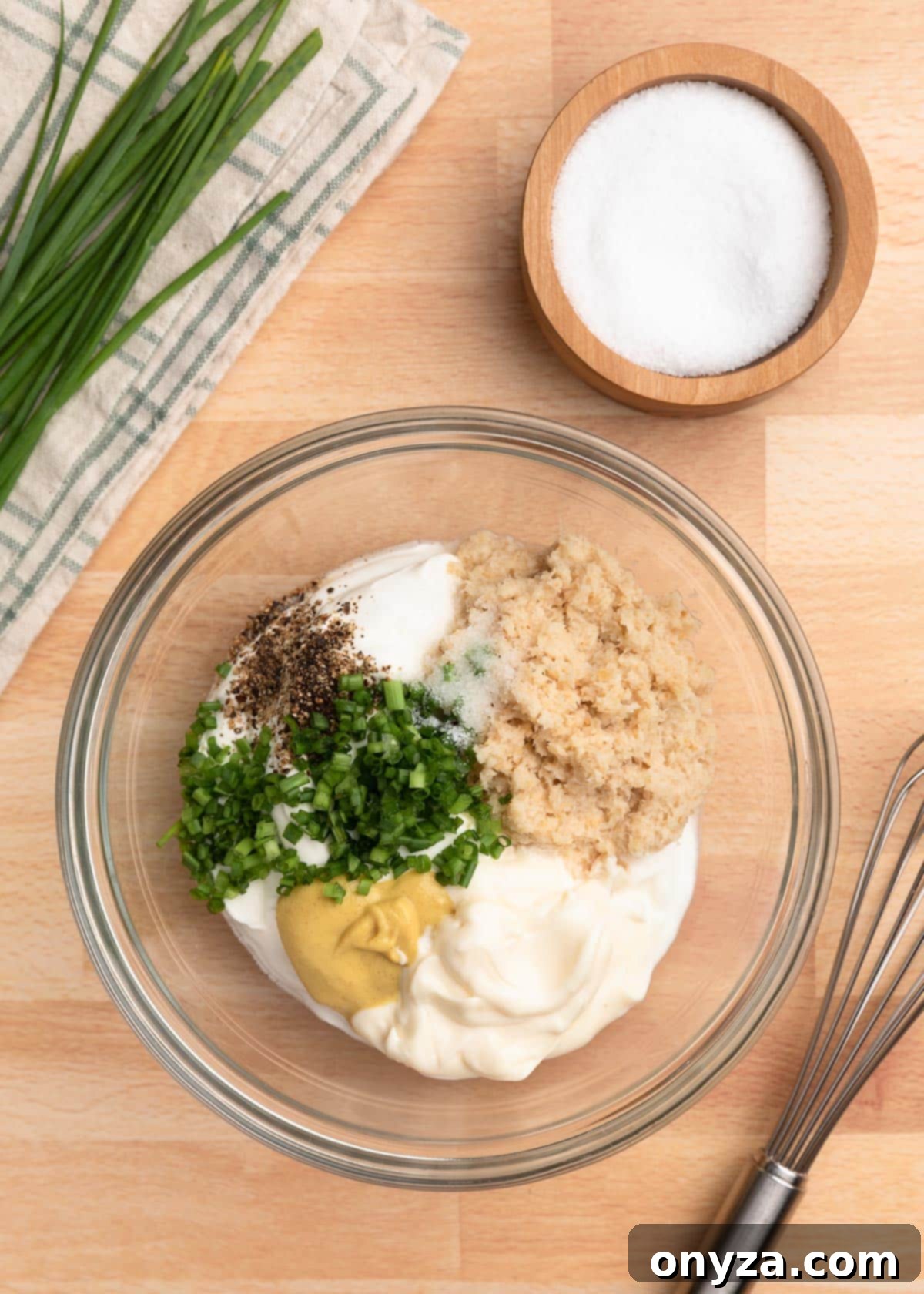 overhead photo of ingredients for creamy horseradish sauce in a mixing bowl, with a whisk and wooden bowl of salt on the side