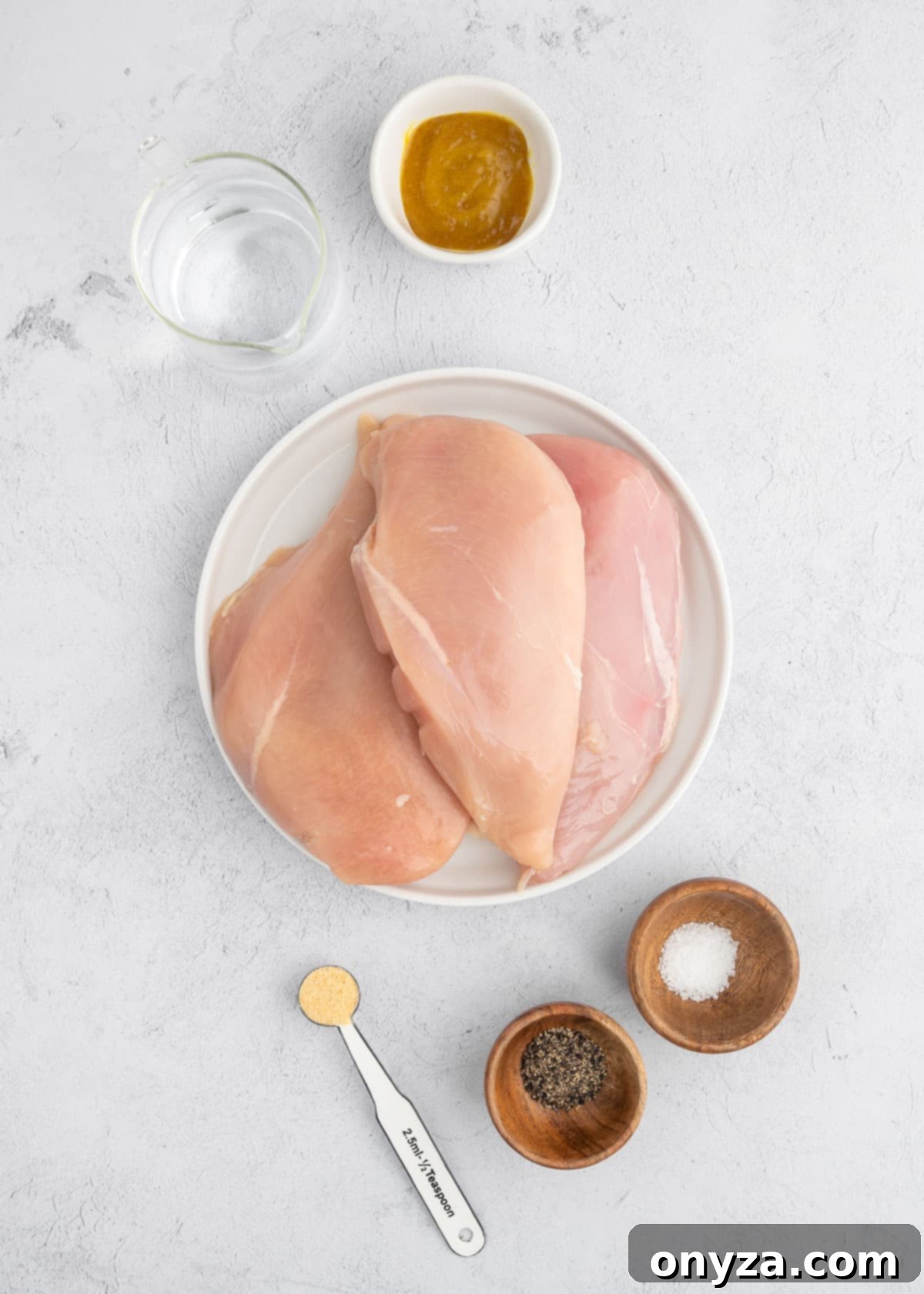 raw chicken breasts on a white plate surrounded small bowls and measuring cups of water, chicken base, garlic powder, pepper, and salt