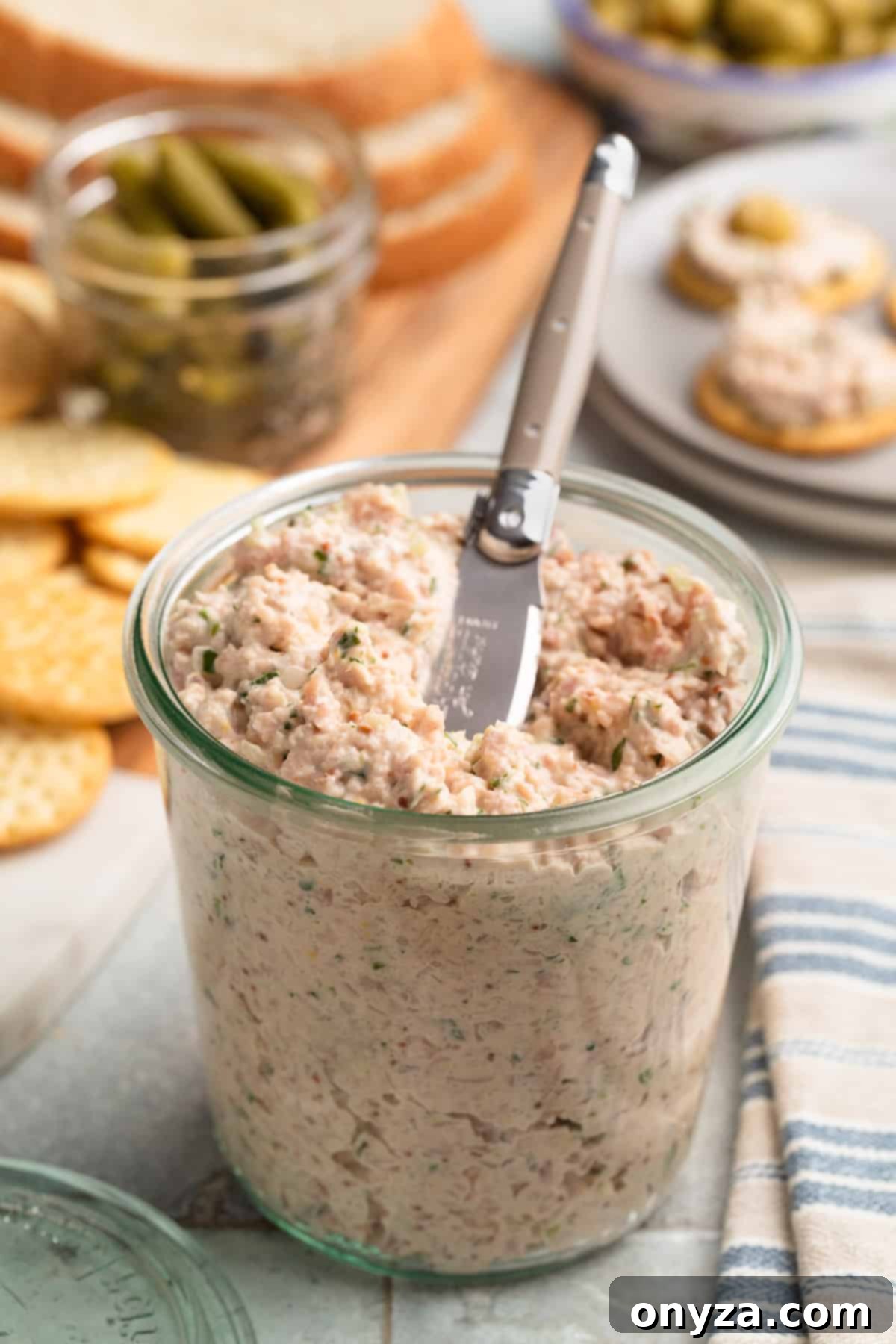 Deviled ham spread in a glass jar with a spreader, in front of a board and plate with crackers, cornichons, and slices of rye bread, showcasing a rich and inviting texture.