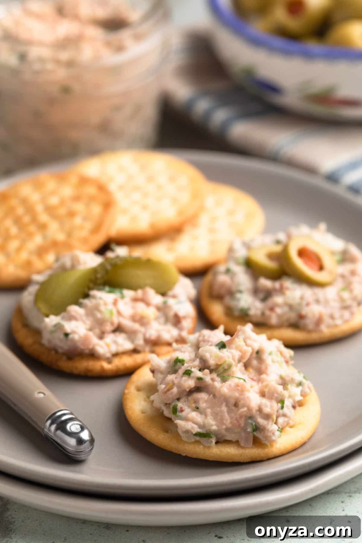 Deviled ham spread generously applied to various crackers on a plate. One cracker is plain, another is topped with a sliced cornichon, and a third is adorned with a sliced pimento-stuffed olive, demonstrating elegant garnishing options.