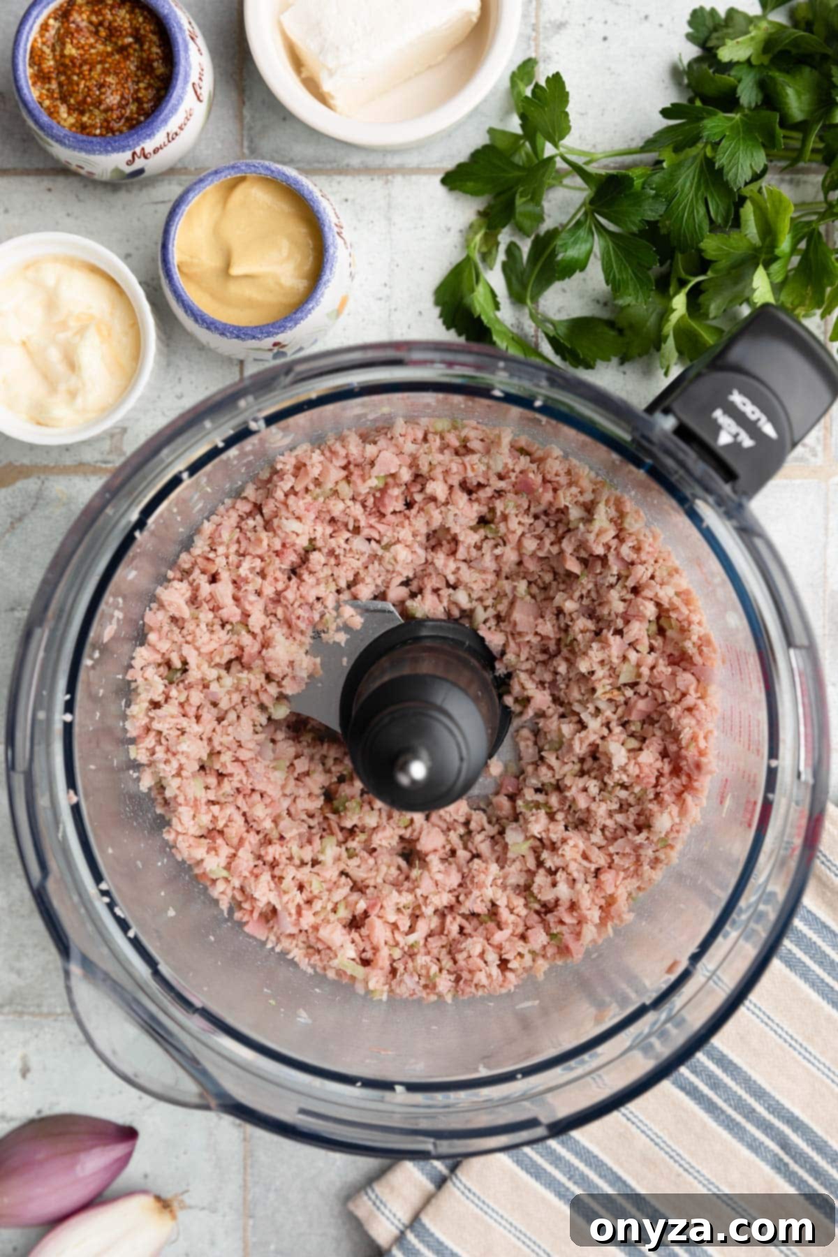 Overhead photo of finely minced ham, celery, and shallots in the bowl of a food processor after pulsing, showing the ideal consistency for the spread.