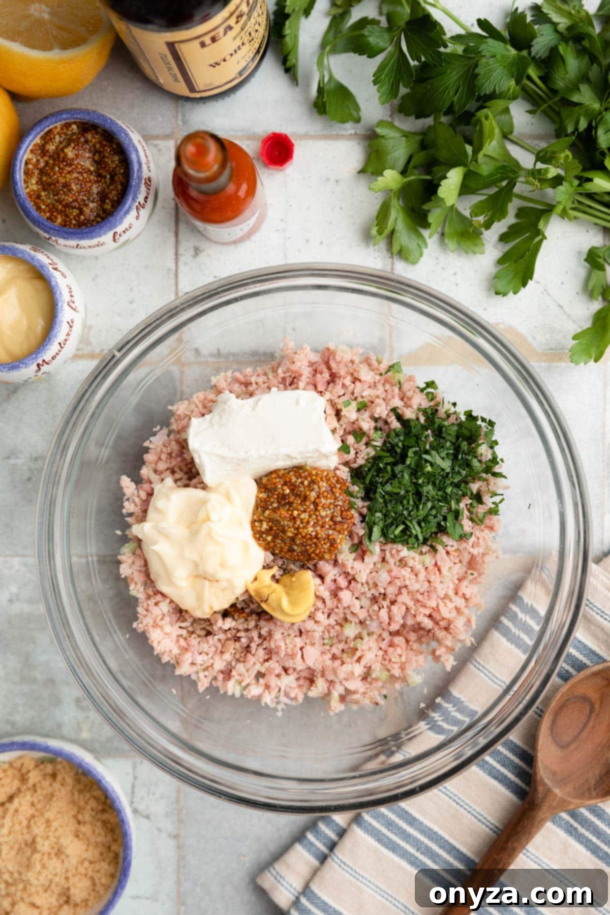 Overhead photo of the minced ham, celery, and onion in a glass bowl, with softened cream cheese, mayonnaise, fresh parsley, and mustards arranged around it, ready for hand mixing.