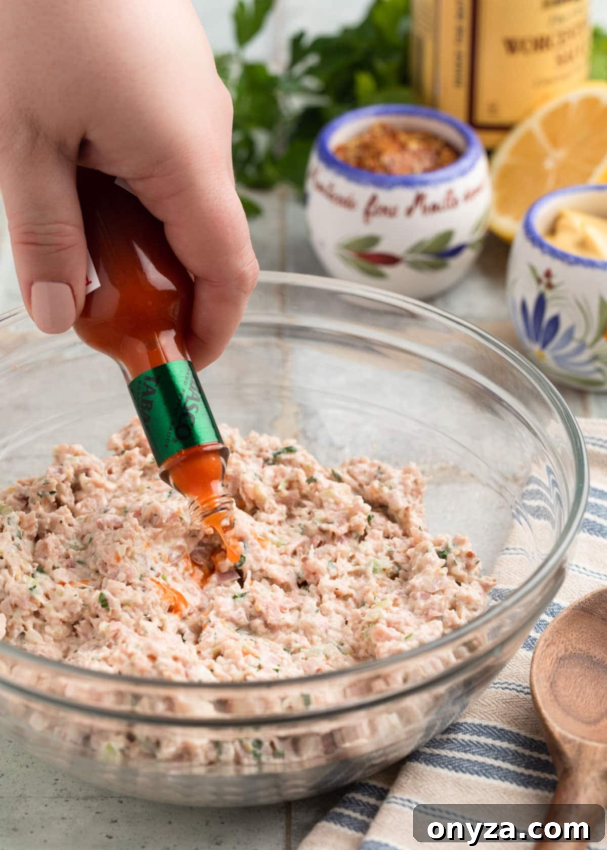 A small glass bottle of hot sauce being shaken into a glass bowl of Deviled Ham spread, illustrating the customization of spiciness.