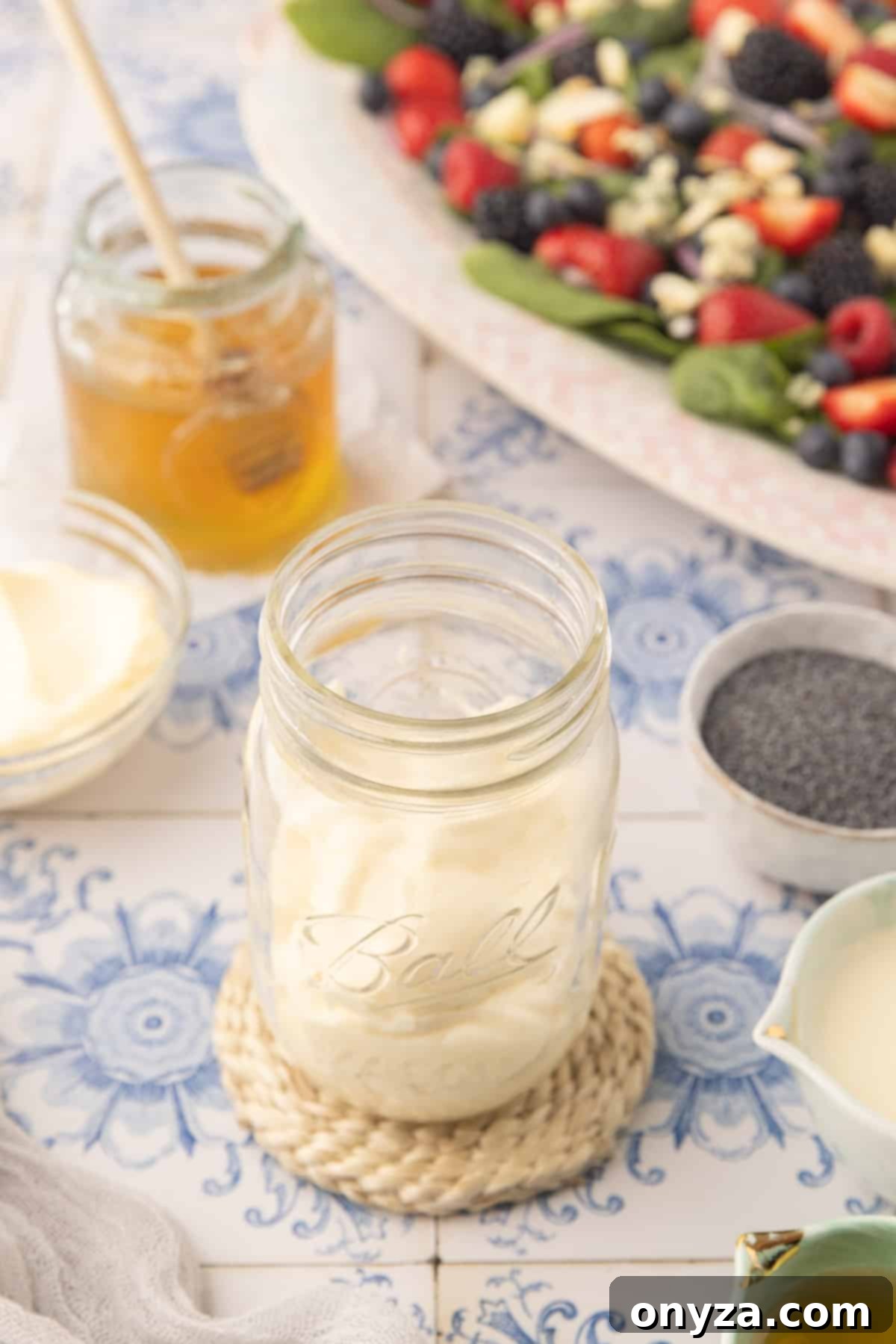 Velvet Poppy Seed Dressing 3 Mayonnaise in a glass mason jar on a blue and white tiled surface, next to a spinach berry salad and a jar of honey.