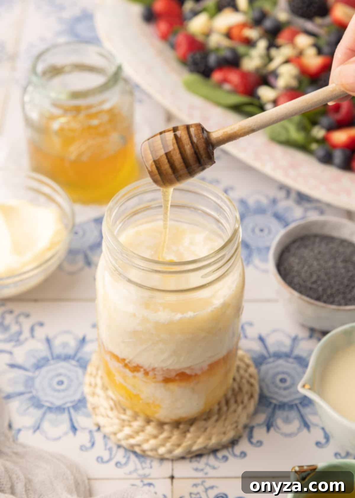 Velvet Poppy Seed Dressing 4 Adding honey from a wooden dipper to a mason jar filled with mayonnaise, apple cider vinegar, and half and half cream, sitting on a blue and white tiled surface.