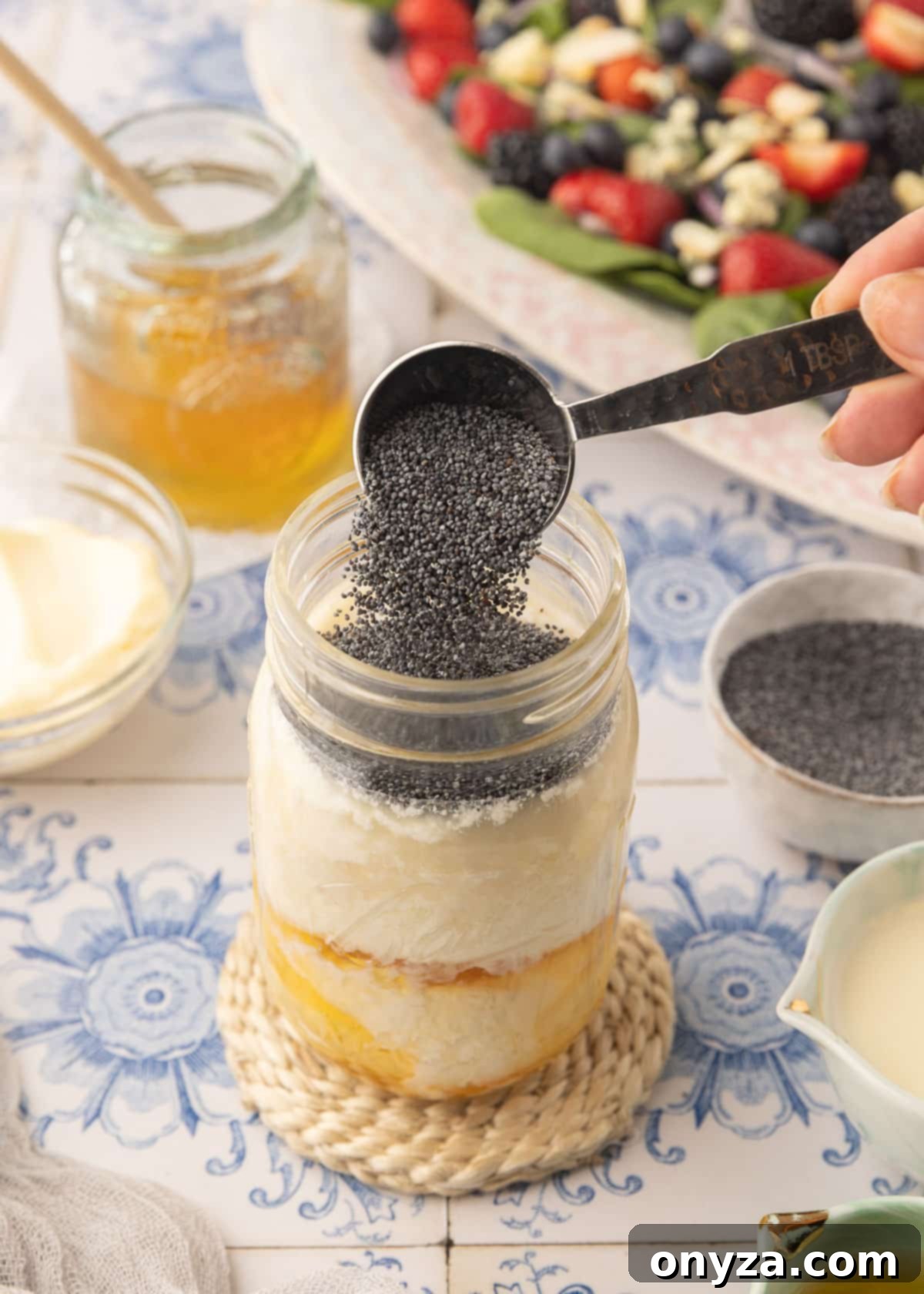 Velvet Poppy Seed Dressing 5 Adding poppy seeds from a tablespoon measure to a mason jar filled with mayonnaise, apple cider vinegar, honey, and half and half cream, sitting on a blue and white tiled surface.