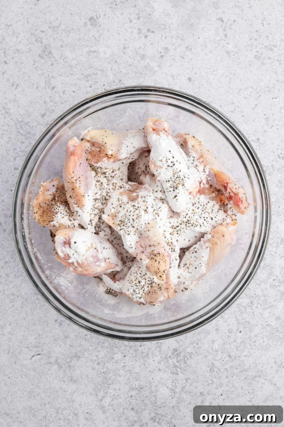 overhead photo of raw split chicken wings in a glass bowl with cornstarch and seasonings sprinkled on top
