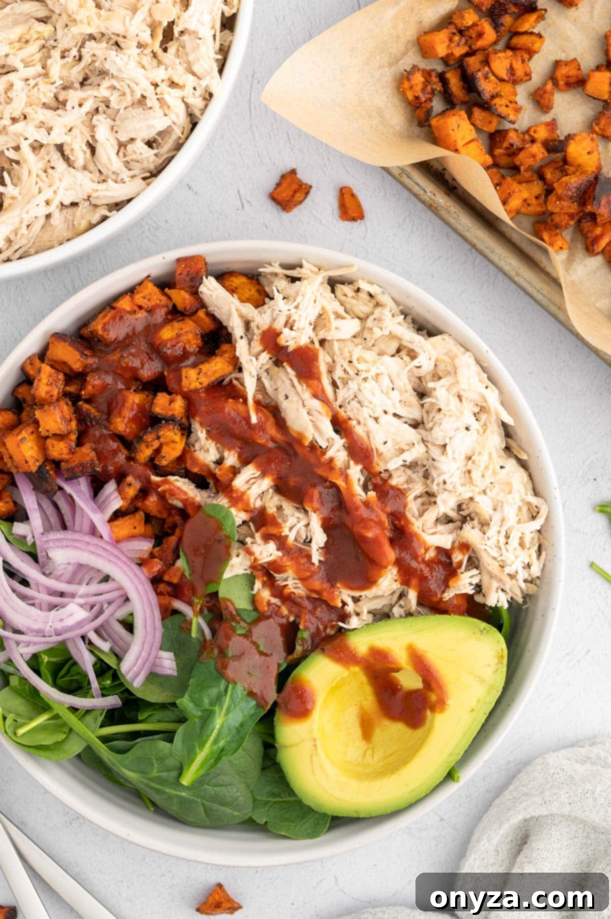 Overhead photo of a vibrant BBQ chicken avocado salad surrounded by a bowl of tender shredded chicken and a sheet pan of roasted sweet potato cubes, suggesting a healthy meal prep option