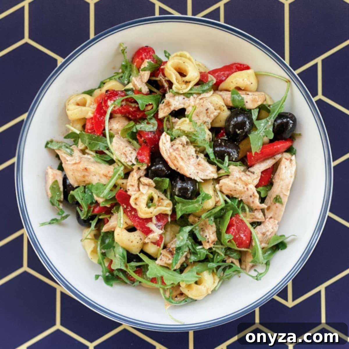 A vibrant chicken tortellini salad in a white bowl, featuring baby arugula, roasted red peppers, and olives, set against a cheerful yellow and blue geometric background