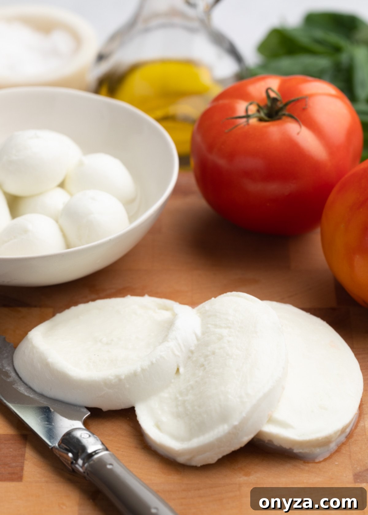 Fresh Caprese Salad 5 sliced buffalo mozzarella on a wood cutting board with tomatoes and a white bowl of ciliegine mozzarella balls.