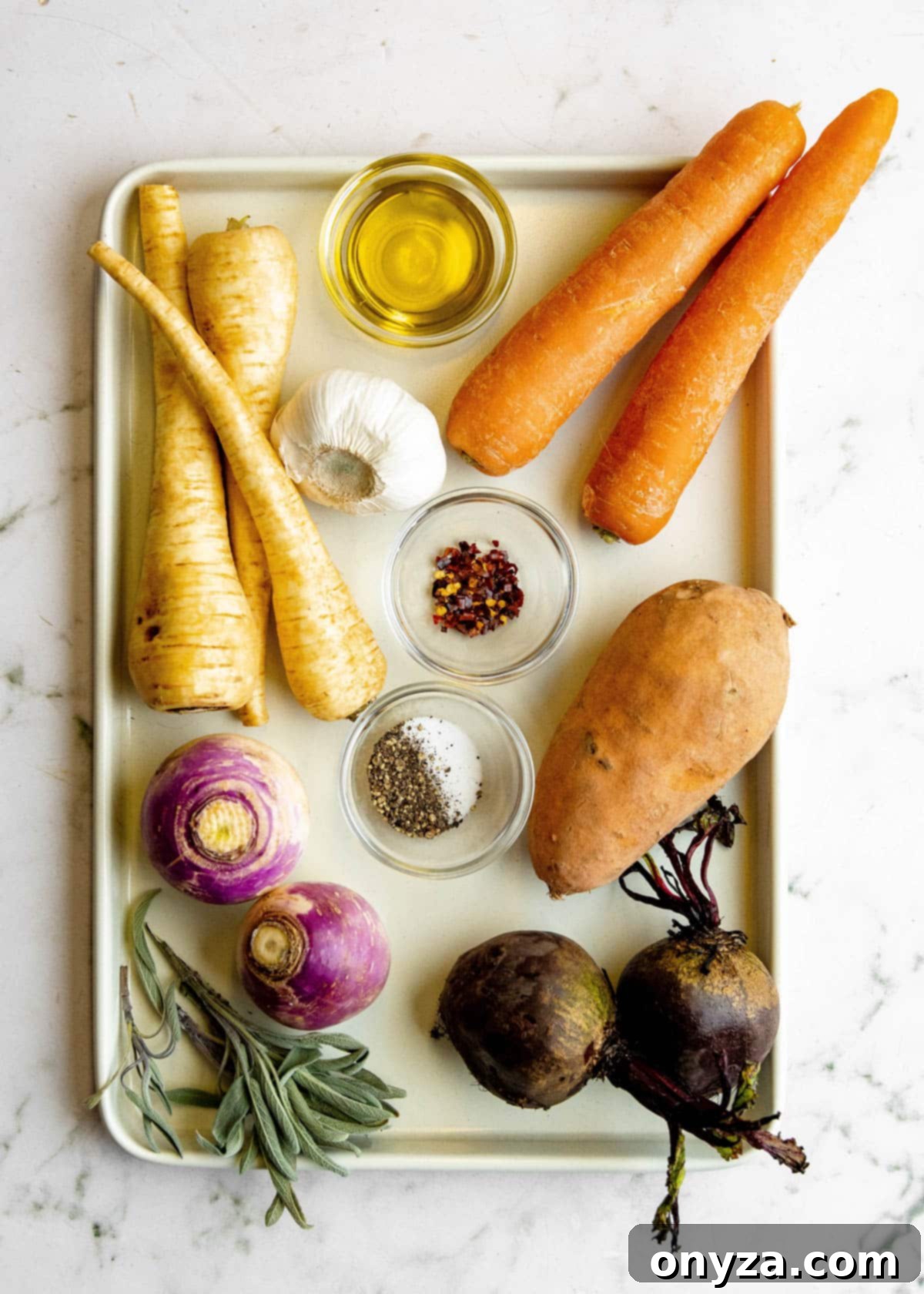 Overhead view of various raw root vegetables neatly arranged on a baking sheet, with small bowls of olive oil and seasonings nearby.