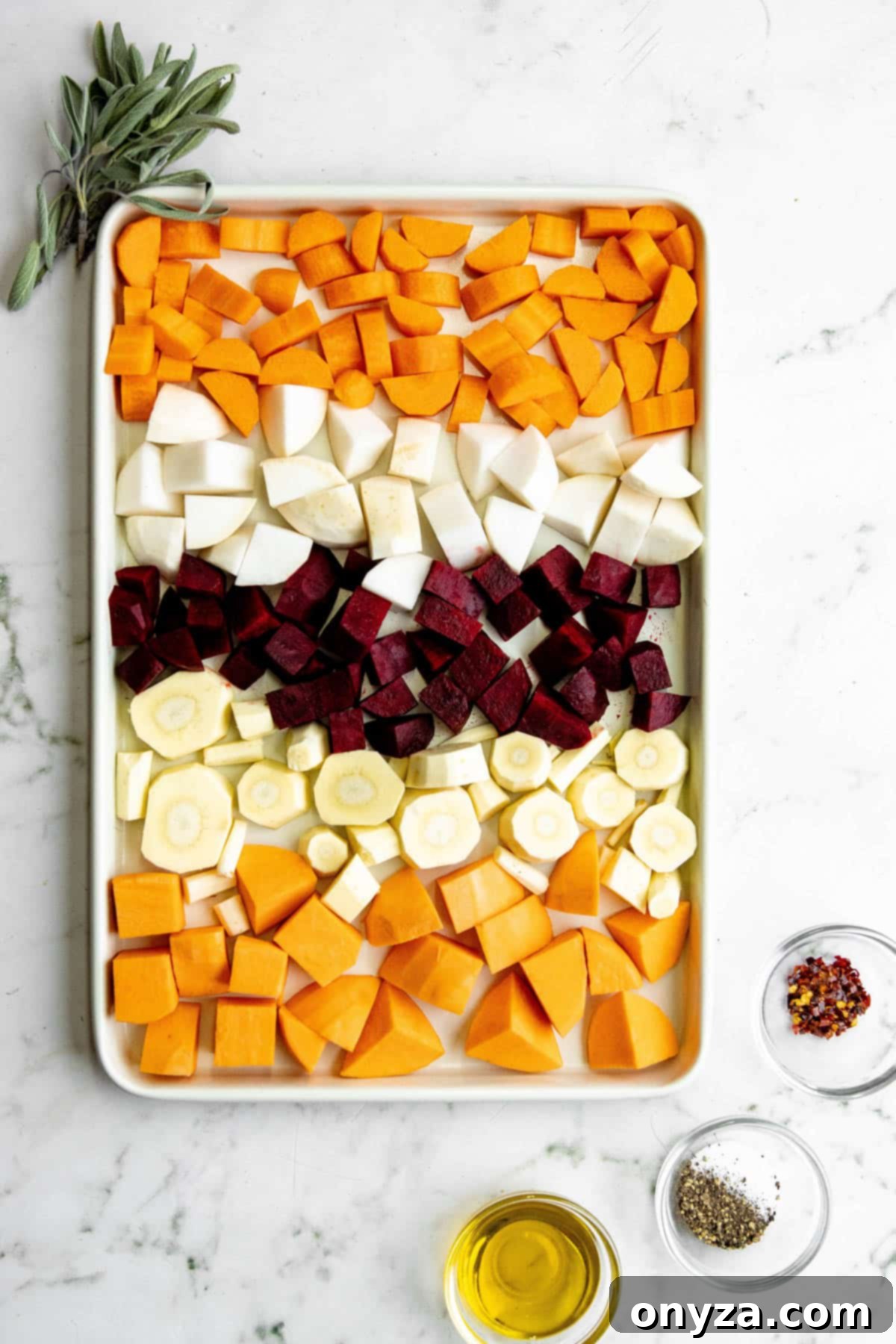 Close-up overhead shot of cubed root vegetables, including carrots, potatoes, and beets, on a baking sheet, with fresh sage sprigs and small bowls of olive oil and spices.