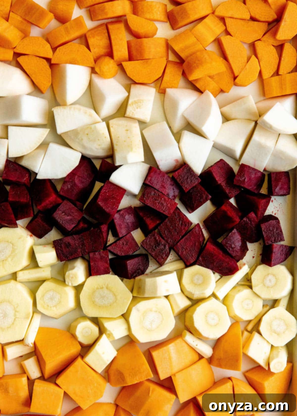 A close-up view of uniformly cubed root vegetables, including various types and colors, neatly spread out on a nonstick rimmed baking sheet.