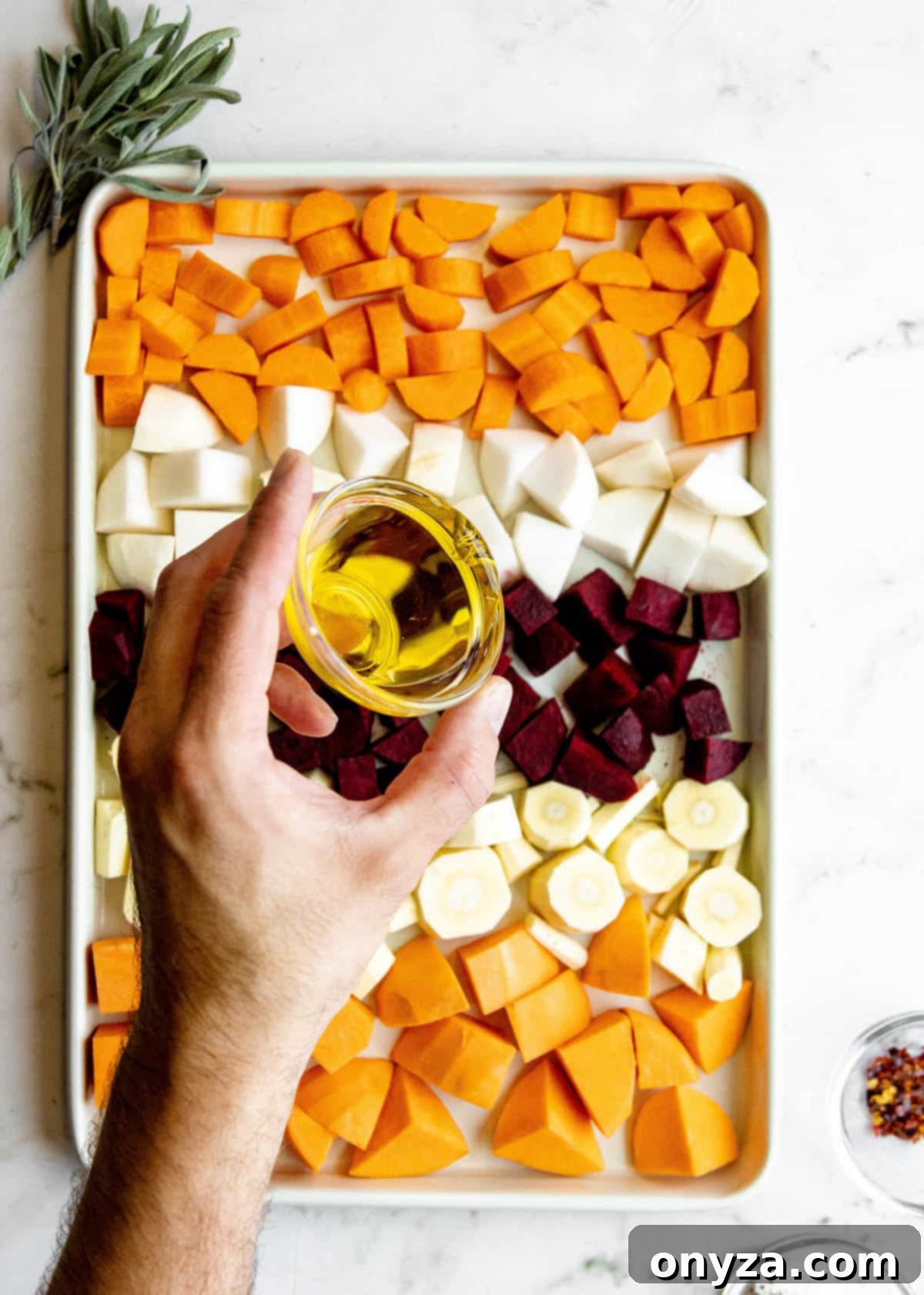 Overhead shot of olive oil being generously drizzled over a vibrant assortment of cubed root vegetables spread across a baking sheet.