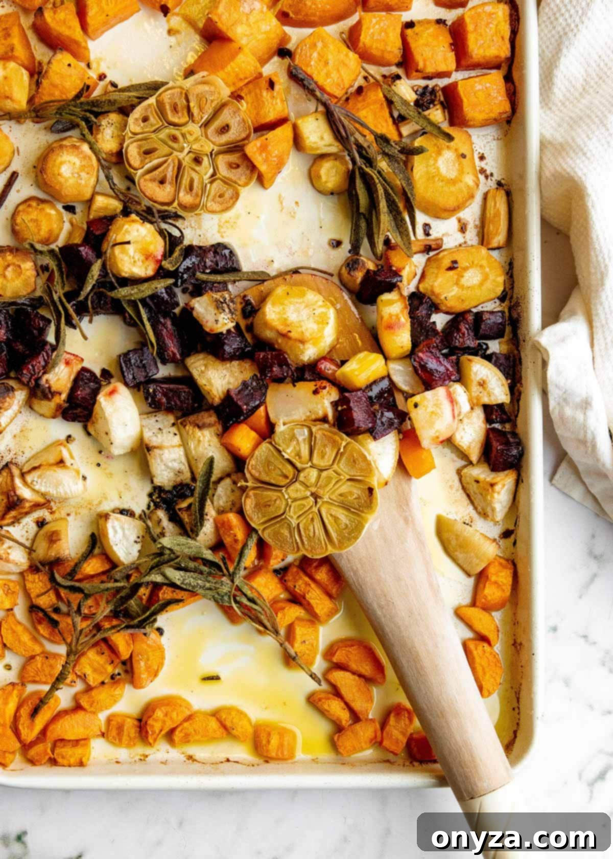 Close-up overhead shot of roasted root vegetables, including colorful carrots, potatoes, and beets, mixed with crispy sage leaves on a baking sheet, with a wooden spoon resting nearby.