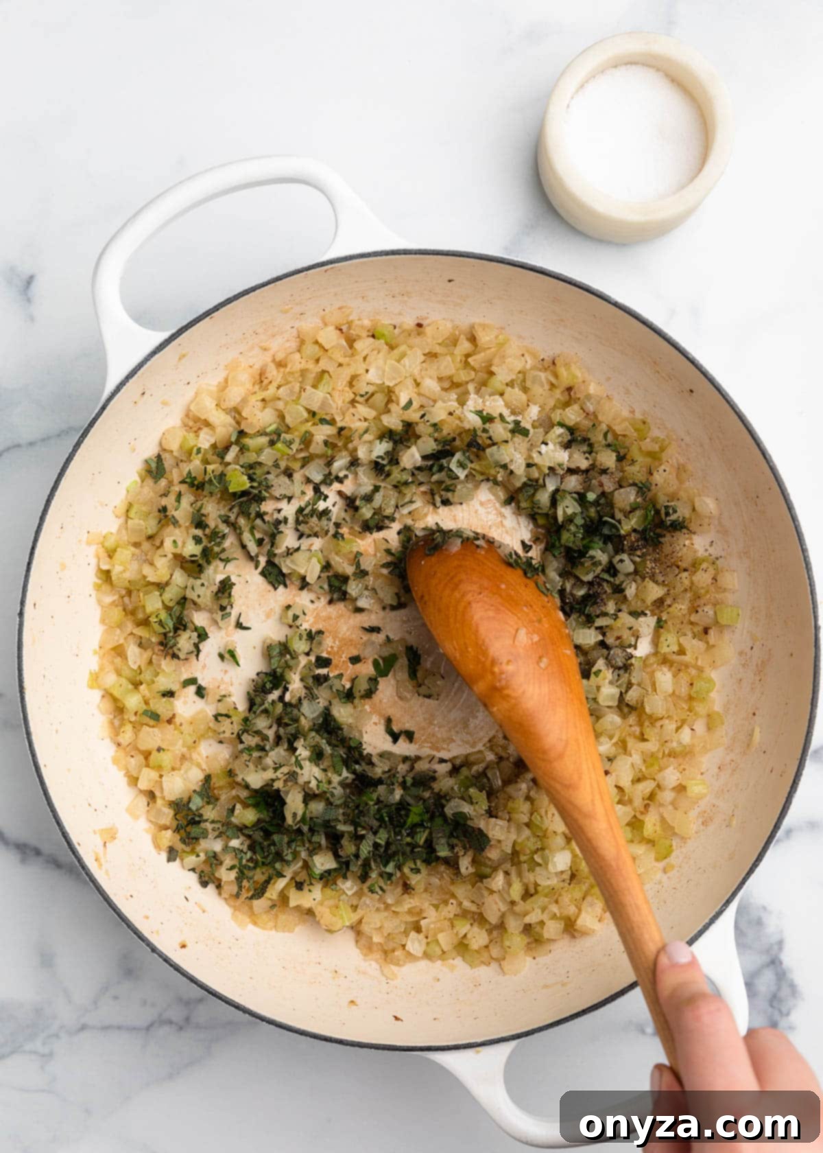 Freshly chopped herbs and seasonings being stirred into cooked celery and onions in a white enameled cast iron Dutch oven with a wooden spoon.