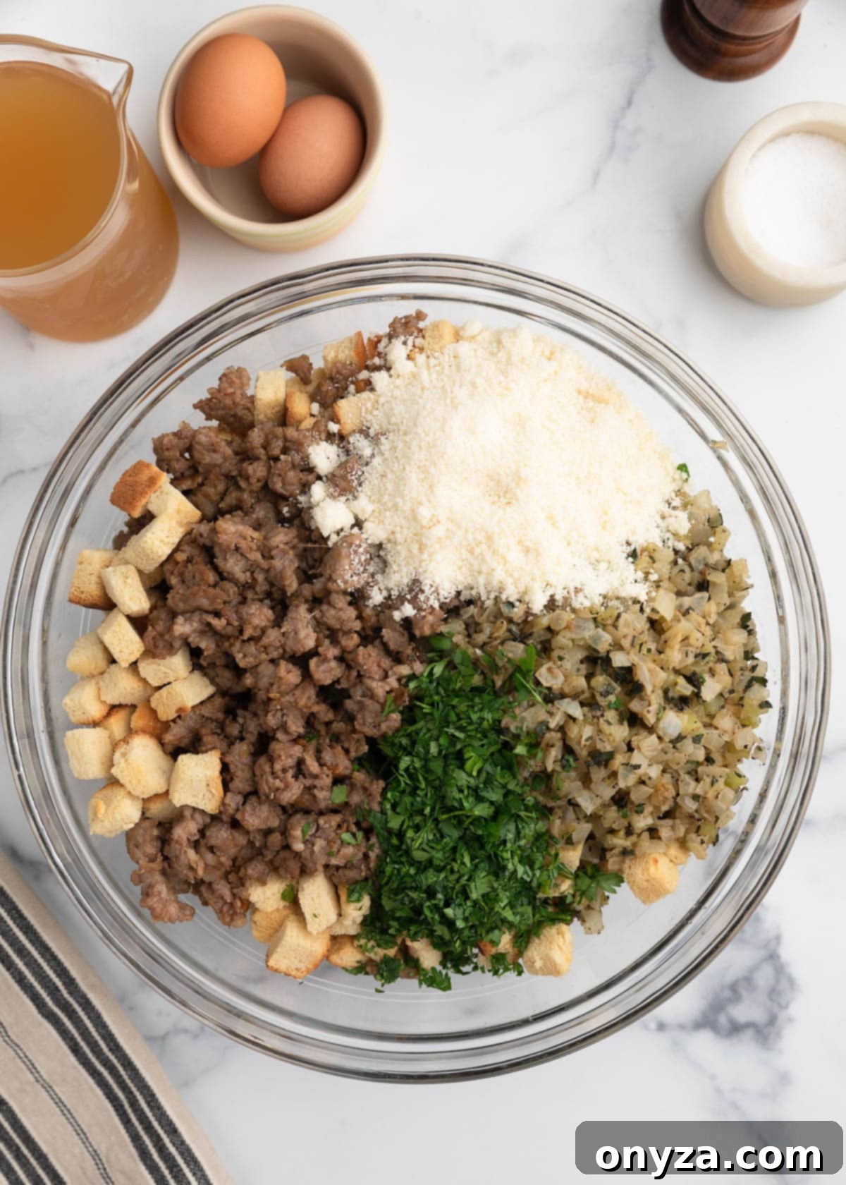 Overhead view of a glass mixing bowl filled with stuffing ingredients, including cooked Italian sausage, cubed bread, sautéed onions and celery, chopped parsley, and grated cheese, surrounded by eggs, broth, and seasonings on a marble surface.