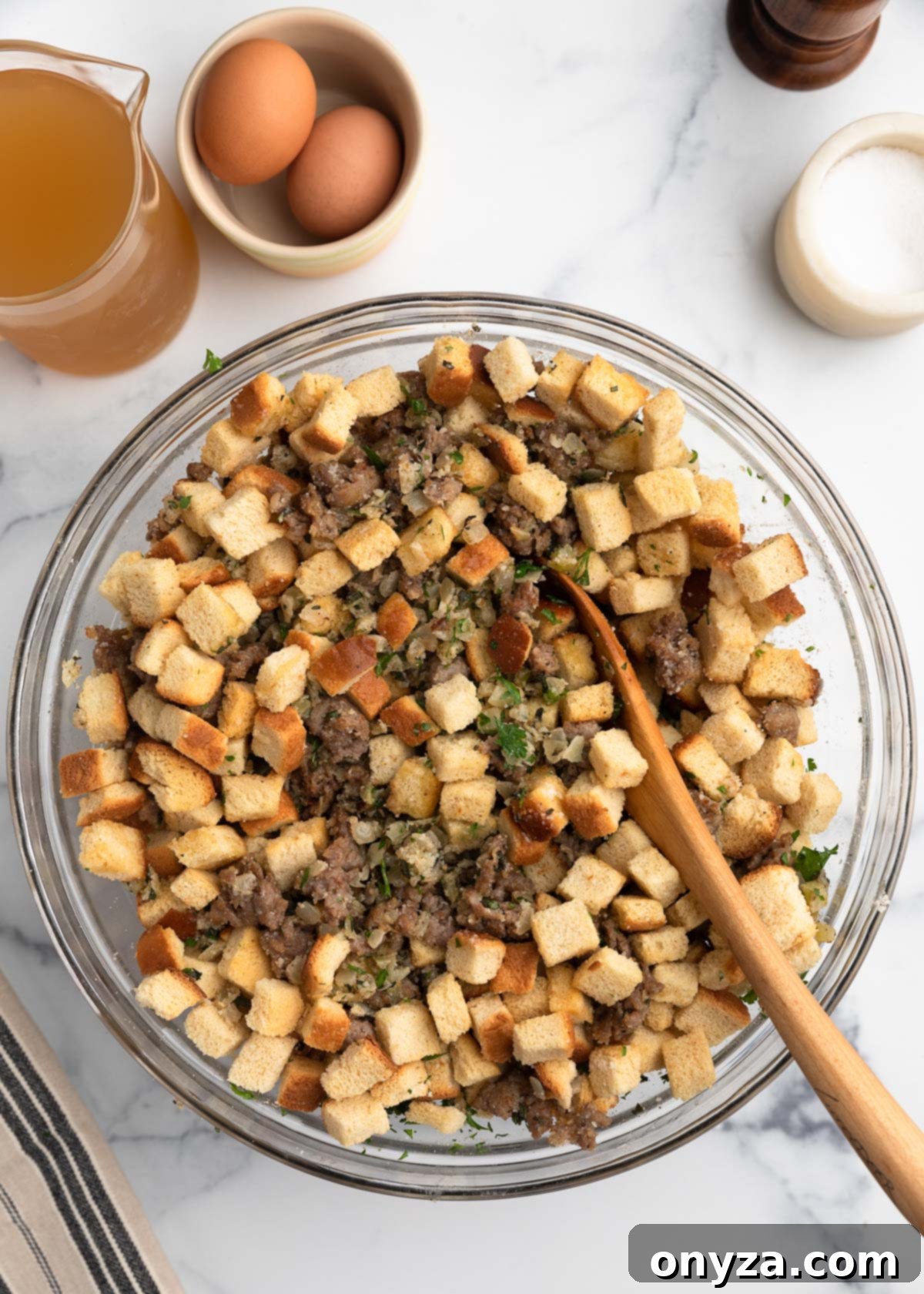 Overhead view of partially mixed Italian sausage stuffing in a glass bowl, showing cubed bread, sausage, herbs, and vegetables being combined with a wooden spoon, with eggs and broth nearby on the countertop.