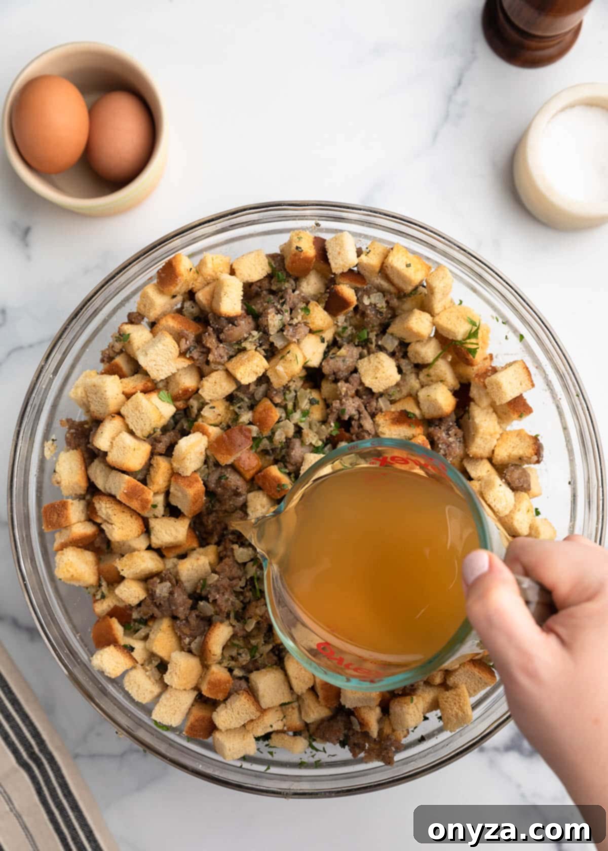A hand pouring turkey stock from a glass measuring cup into a large glass bowl filled with cubed bread, cooked sausage, onions, and fresh herbs for stuffing.