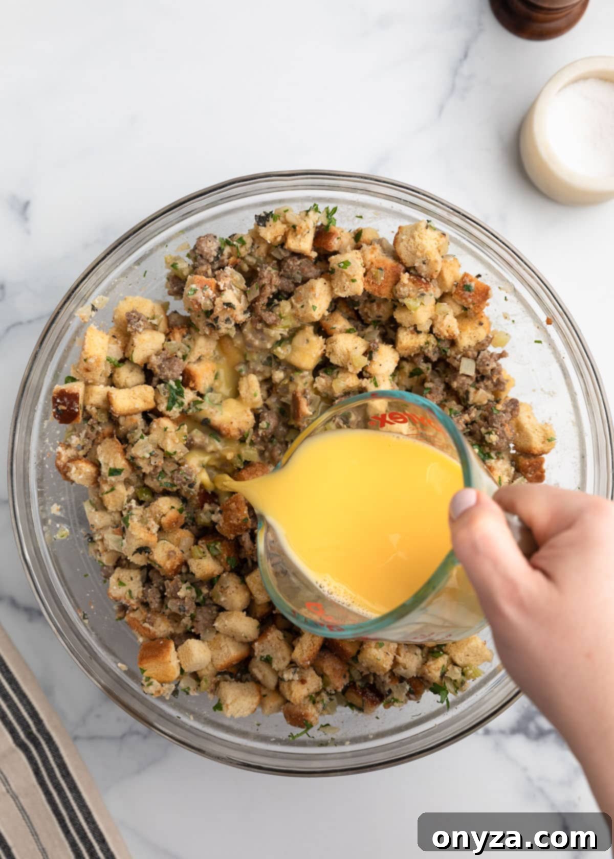 Beaten eggs and stock being poured from a measuring cup into a bowl of sausage stuffing mixture with bread cubes, herbs, and broth on a white marble surface.