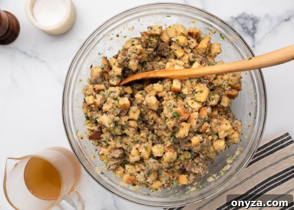 Overhead view of Italian sausage stuffing mixture in a large glass mixing bowl with a wooden spoon. Chopped bread, sausage, celery, and herbs are visible, and a pitcher of broth sits beside the bowl, indicating readiness to add more moisture if needed.