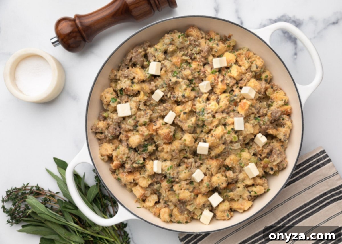 Overhead view of unbaked Italian sausage stuffing in a white braiser, dotted with small cubes of butter on top. Fresh herbs, salt, and pepper are arranged nearby on a marble surface.