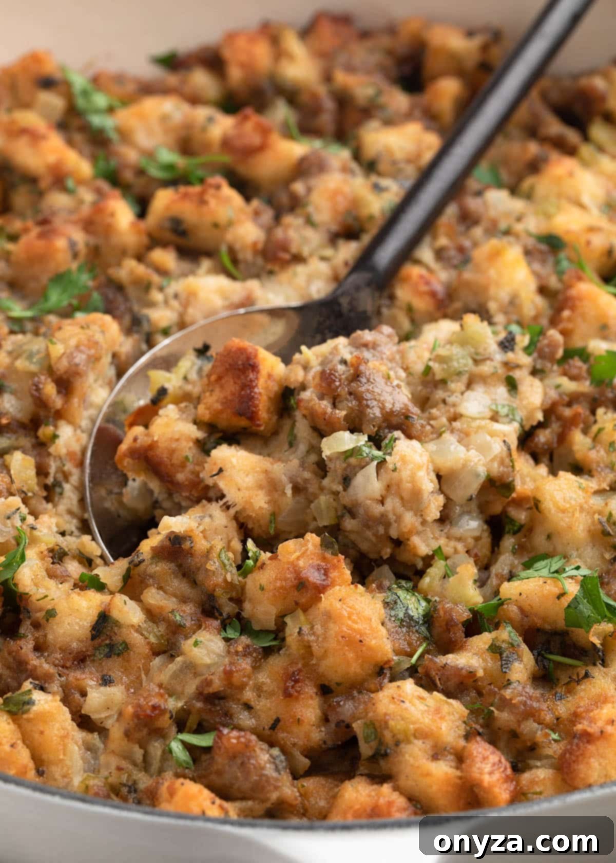 Close-up of a serving spoon scooping Italian sausage stuffing from a white dish, showcasing browned bread cubes, crumbled sausage, onions, celery, and chopped parsley.