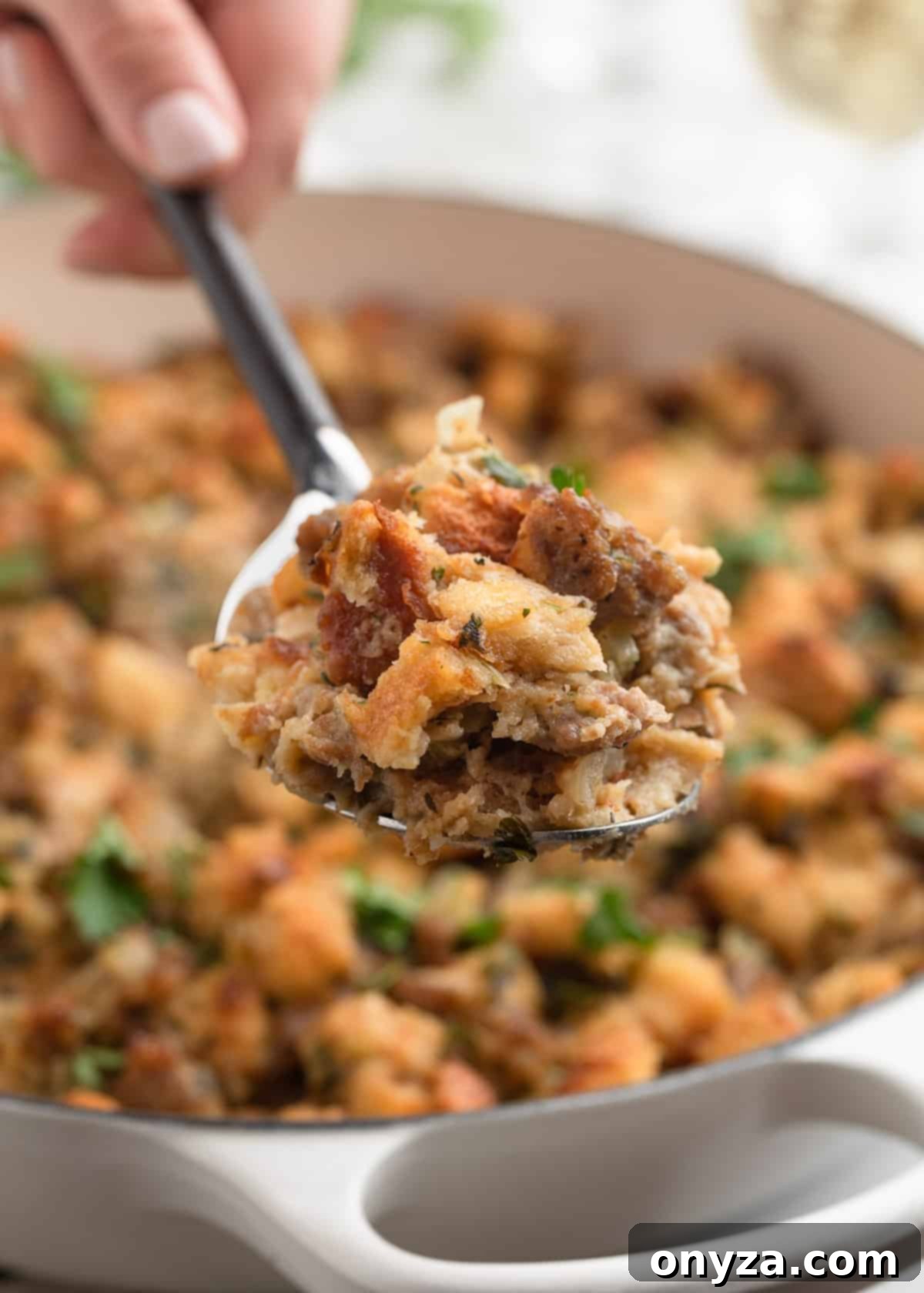 A close-up of a spoonful of baked Italian sausage stuffing being lifted from a white casserole dish, showing golden toasted bread cubes, browned sausage, and chopped herbs, emphasizing its fluffy and flavorful texture.