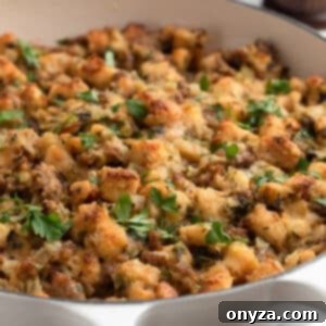 Golden-brown Italian sausage stuffing in a white braiser, topped with chopped fresh parsley. The stuffing features toasted bread cubes, crumbled sausage, and visible bits of onion and celery.