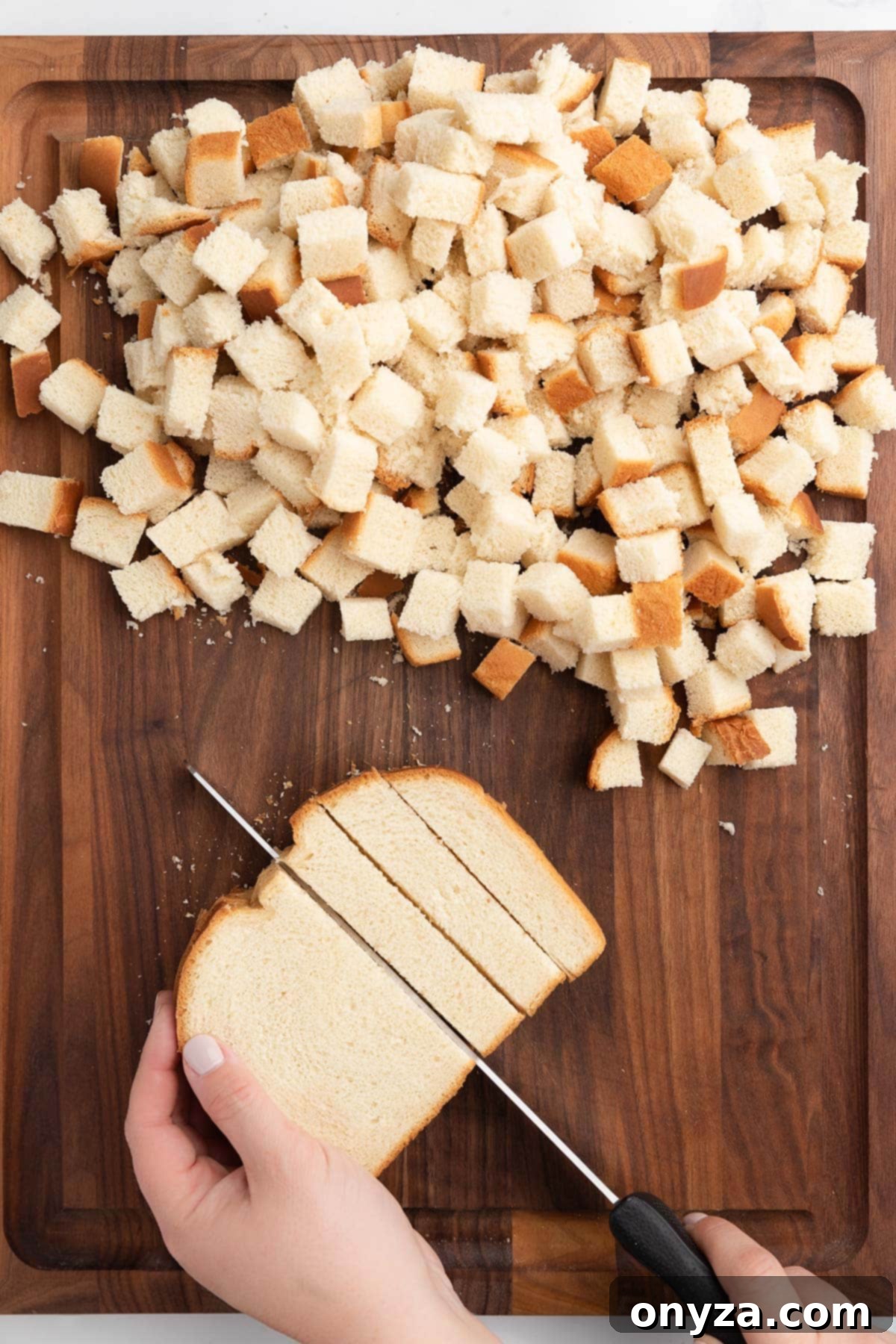 A hand cutting country white bread into even strips with a serrated knife on a wooden cutting board.