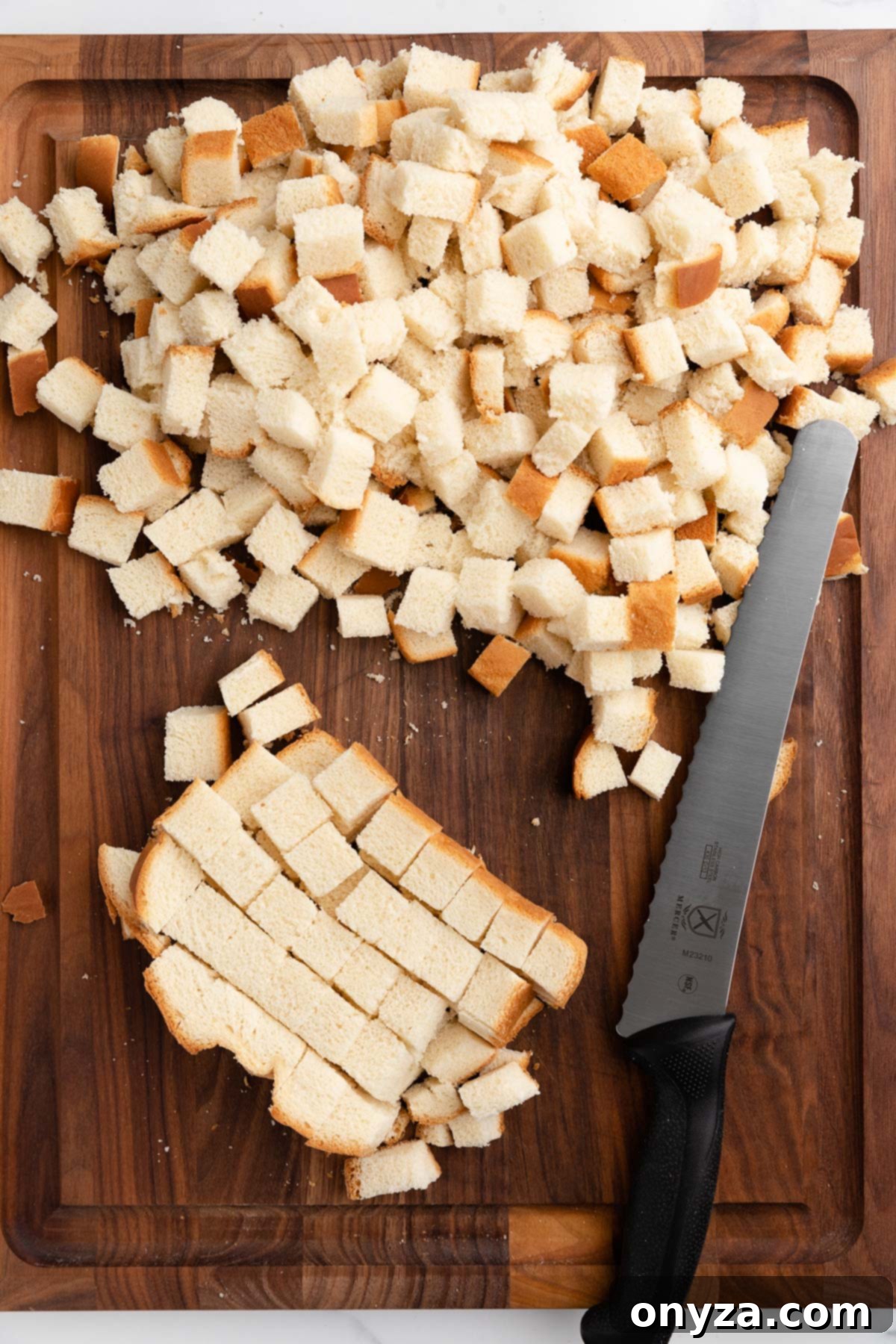 A hand continuing to cut bread strips into uniform cubes with a serrated knife.