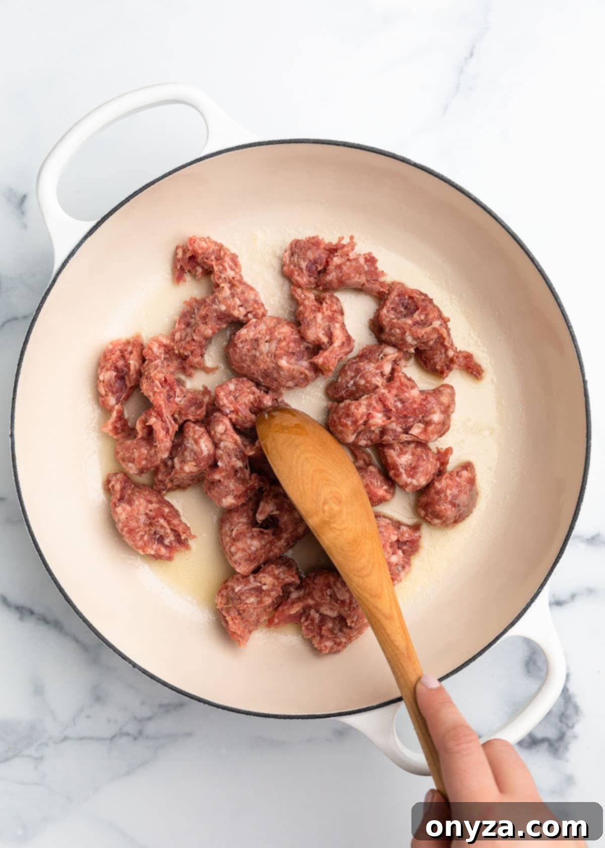 Raw loose Italian sausage meat being broken up with a wooden spoon in a white enameled cast iron Dutch oven.