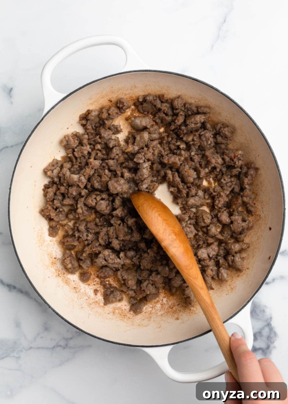 Perfectly browned Italian sausage crumbles in a white enameled cast iron Dutch oven with a wooden spoon.
